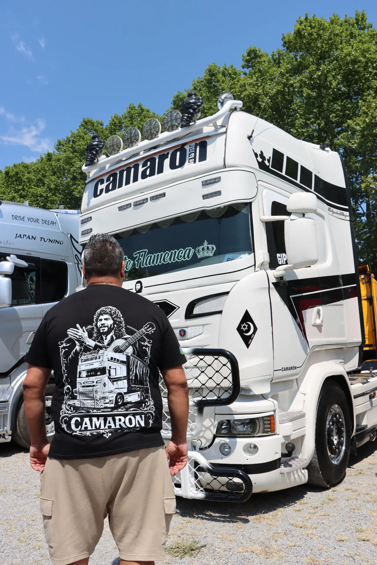 Un hombre con una camiseta de Camaron posa delante de un camión blanco decorado con la palabra «Camaron» en el parabrisas, en la concentración de camiones de Olot.