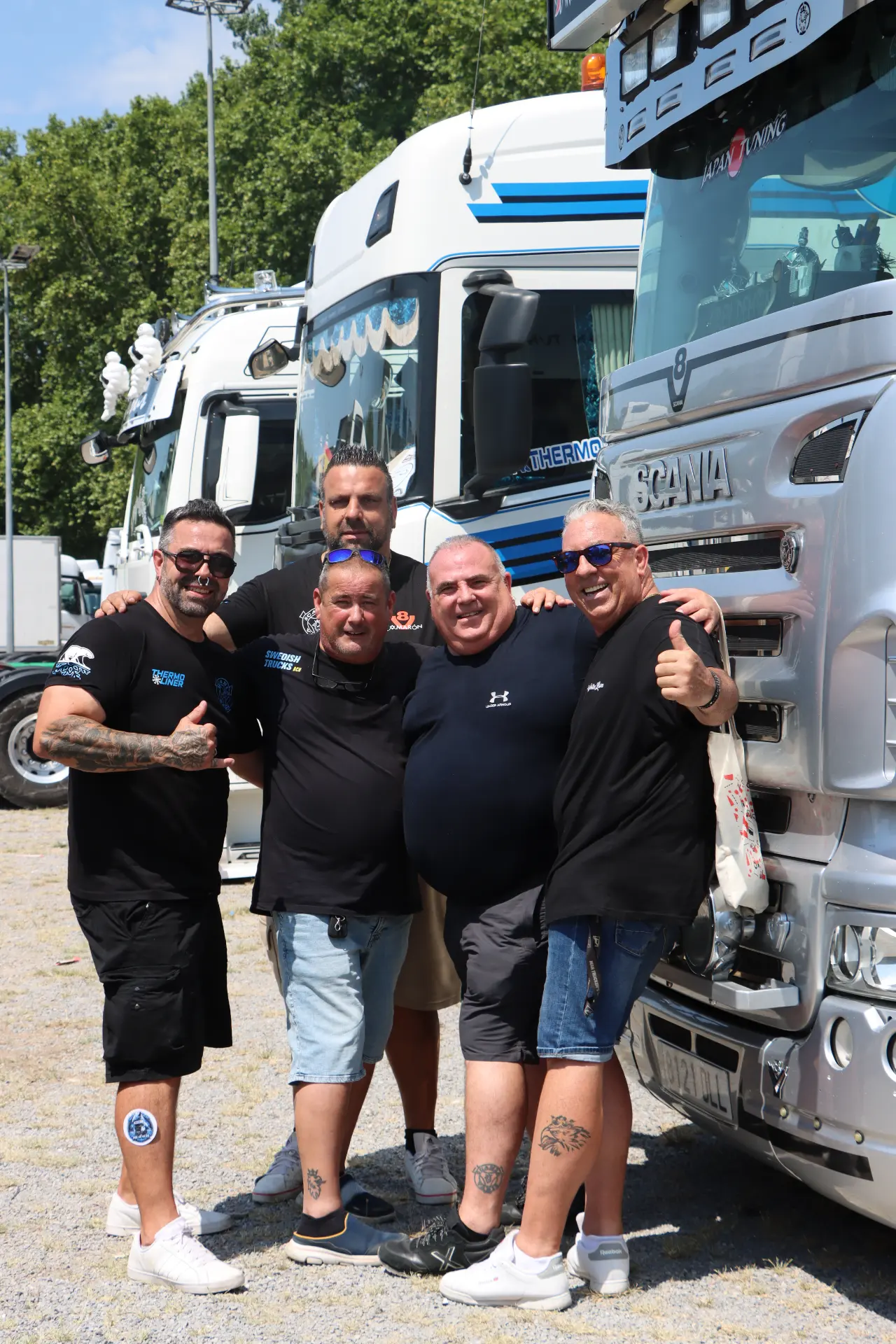 Cinco hombres sonrientes del equipo de Olot posan juntos delante de unos camiones grandes en un día soleado, levantando el pulgar.