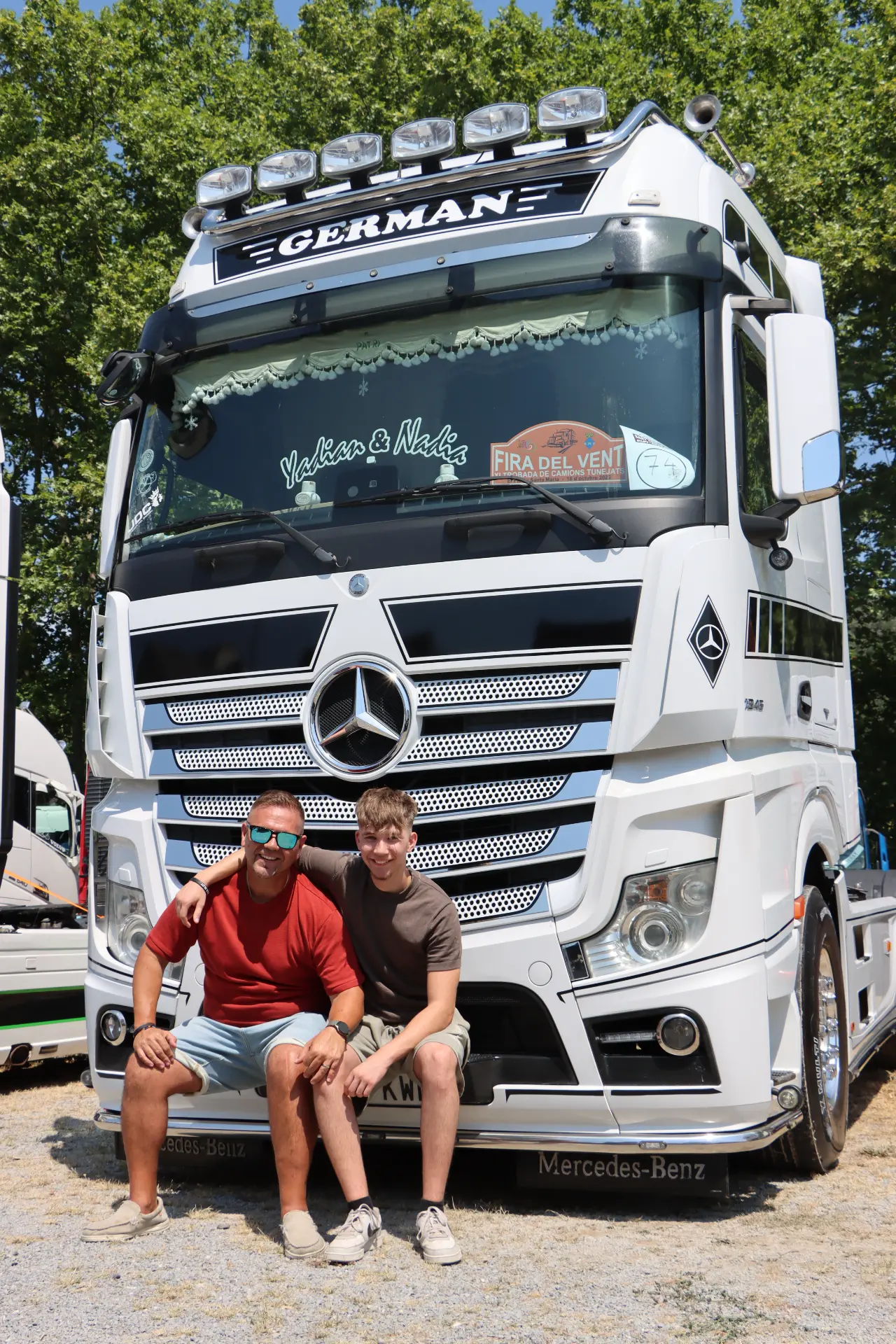 Dos hombres sentados y sonriendo delante de un gran camión Mercedes-Benz blanco aparcado al aire libre en la concentración de camiones de Olot.