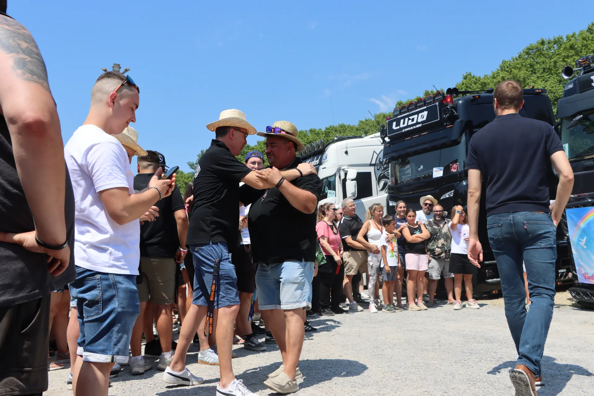 Dos hombres abrazándose y sonriendo entre la multitud, cerca de unos camiones aparcados, en un día soleado durante la concentración de camiones de Olot, un momento especial que reflejó la sincera camaradería del homenaje a Oti Cabadas.