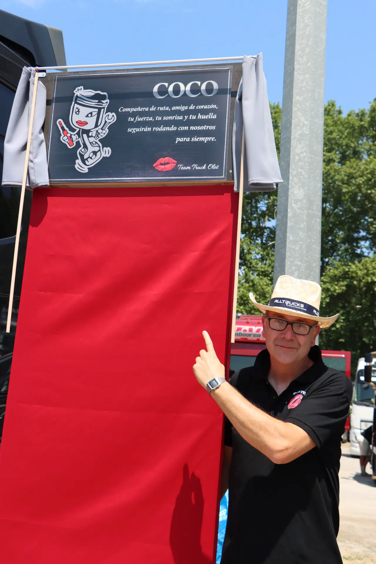 Un hombre con sombrero y gafas señala un cartel rojo en el que se lee «COCO» en el exterior, como miembro del equipo Team Truck Olot durante un homenaje a Oti Cabadas.