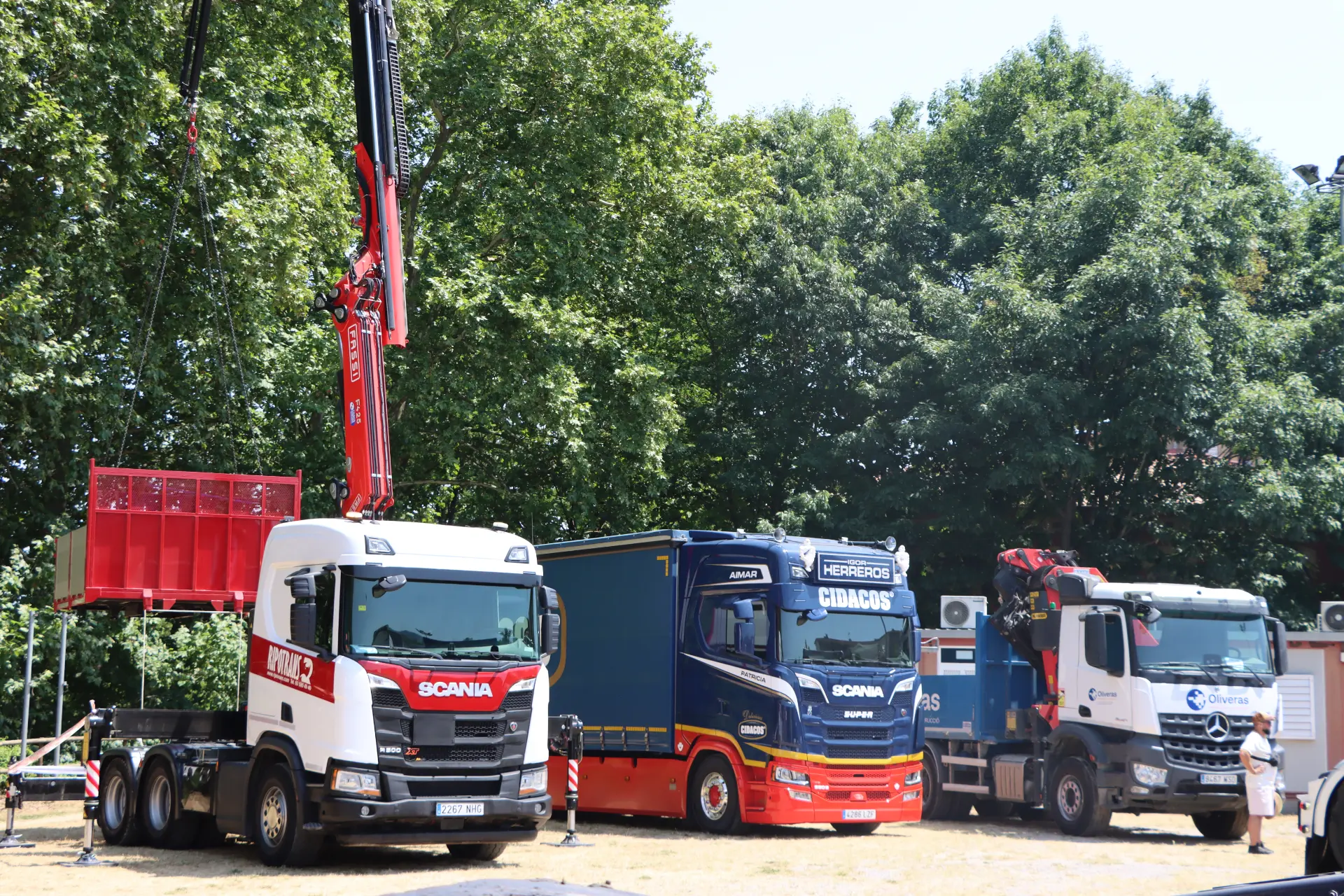 Tres camiones aparcados al aire libre, entre ellos uno del equipo «Team Truck Olot», con una grúa que está levantando un gran contenedor rojo. Árboles al fondo.