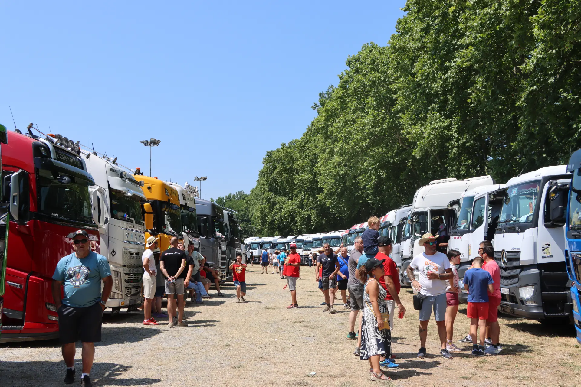 La gente camina entre hileras de camiones aparcados durante la concentración de camiones de Olot, en un día soleado, con árboles a ambos lados.