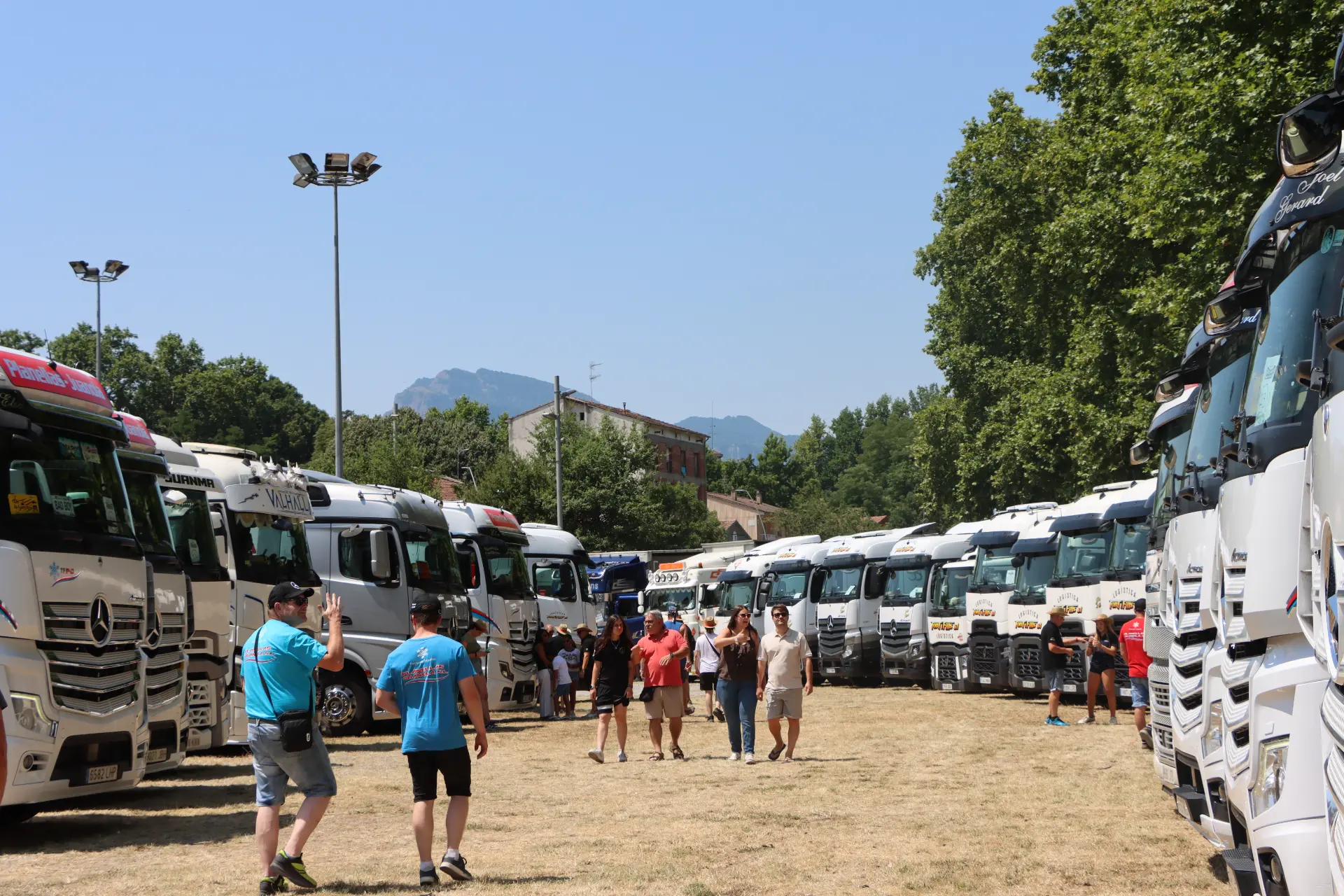 La gente camina entre filas de camiones aparcados en un evento al aire libre en un día soleado, uniéndose a la comunidad de San Cristóbal Camiones en un sincero homenaje a Oti Cabadas, mientras cocotruckergirl comparte el animado ambiente.