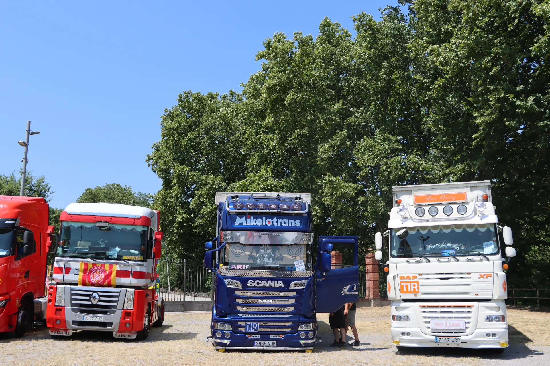 Tres camiones de colores del Team Truck Olot están aparcados uno al lado del otro bajo los árboles, y hay una persona —quizás una «cocotruckergirl»— de pie junto a la puerta del camión del medio, en homenaje a Oti Cabadas.