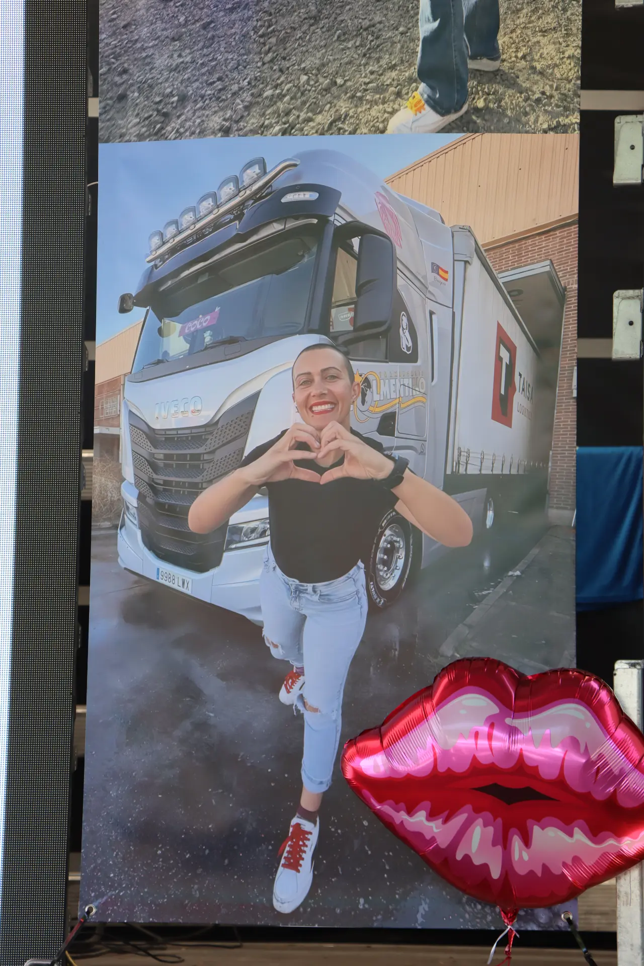 Una persona sonriente forma un corazón con las manos delante de un camión del equipo de Olot; en primer plano, un globo con forma de labios rosas, que refleja el ambiente festivo de la concentración de camiones de Olot.