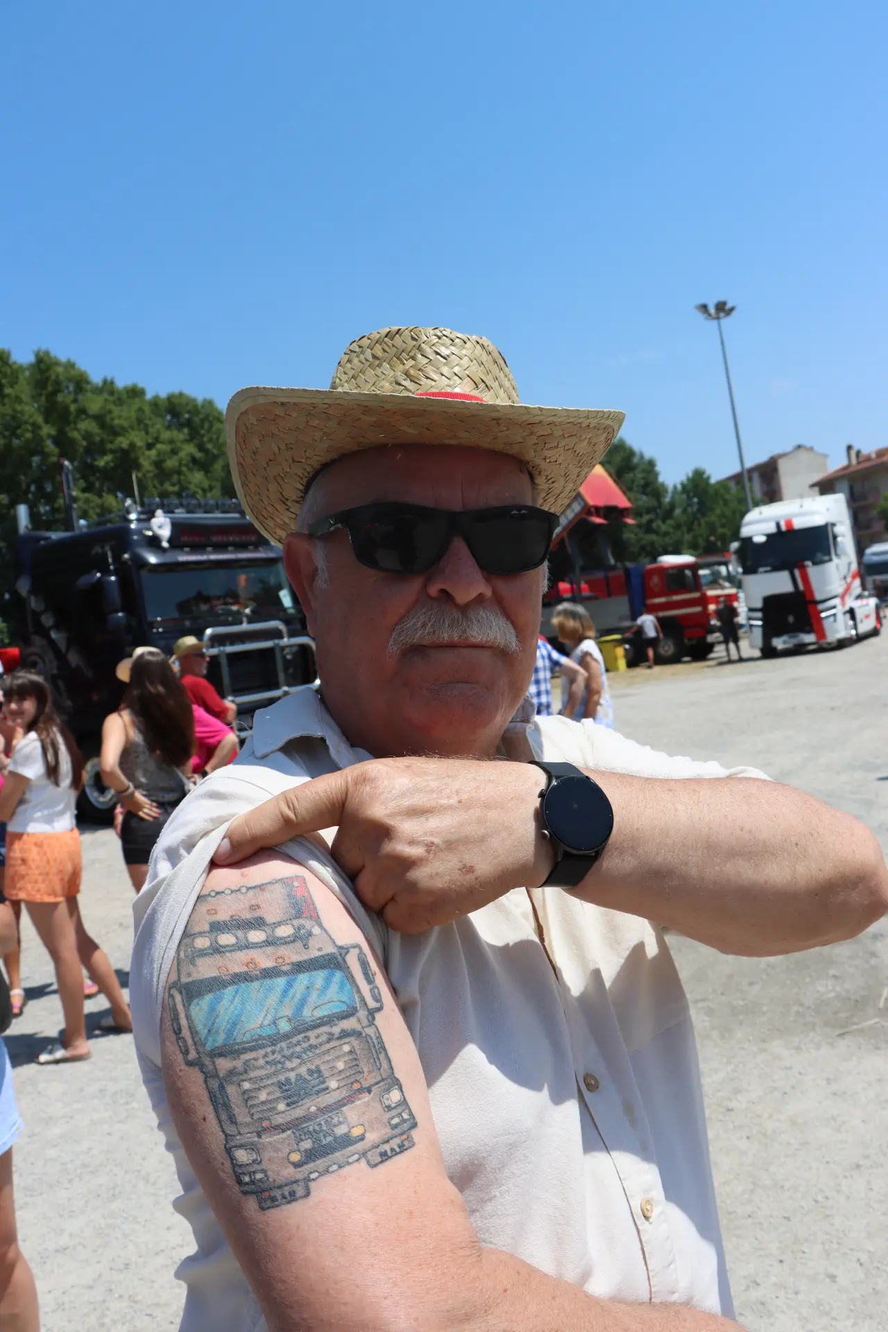 Un hombre con gafas de sol y sombrero de paja señala el tatuaje de un camión que lleva en el brazo durante un acto al aire libre en la «Concentración de Camiones de Olot», con camiones al fondo: un homenaje muy apropiado a Oti Cabadas.