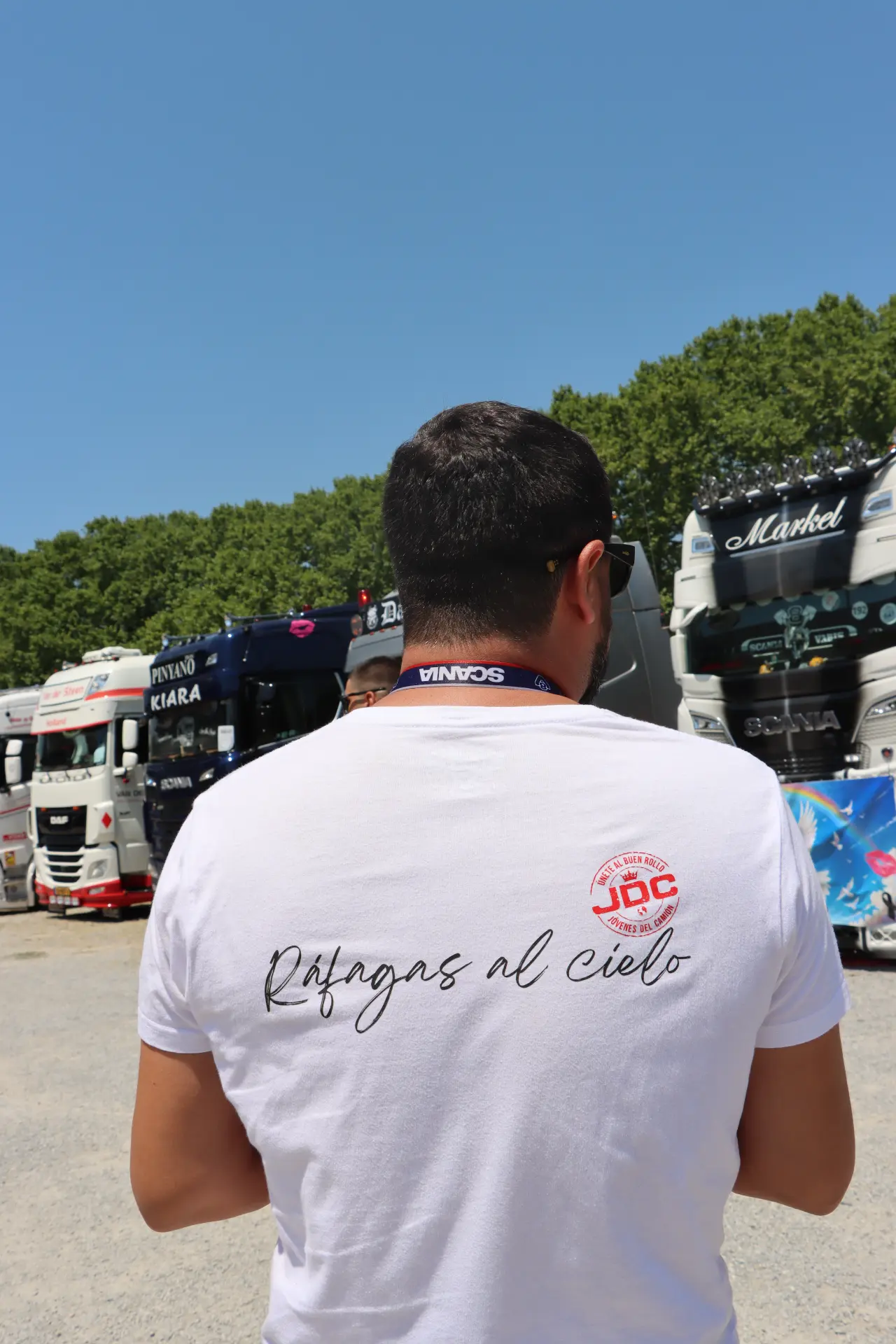 Un hombre con una camiseta blanca con el lema «Ráfagas al cielo» posa delante de unos camiones aparcados bajo un cielo azul y despejado durante la concentración de camiones de Olot, un acto celebrado en homenaje a Oti Cabadas.