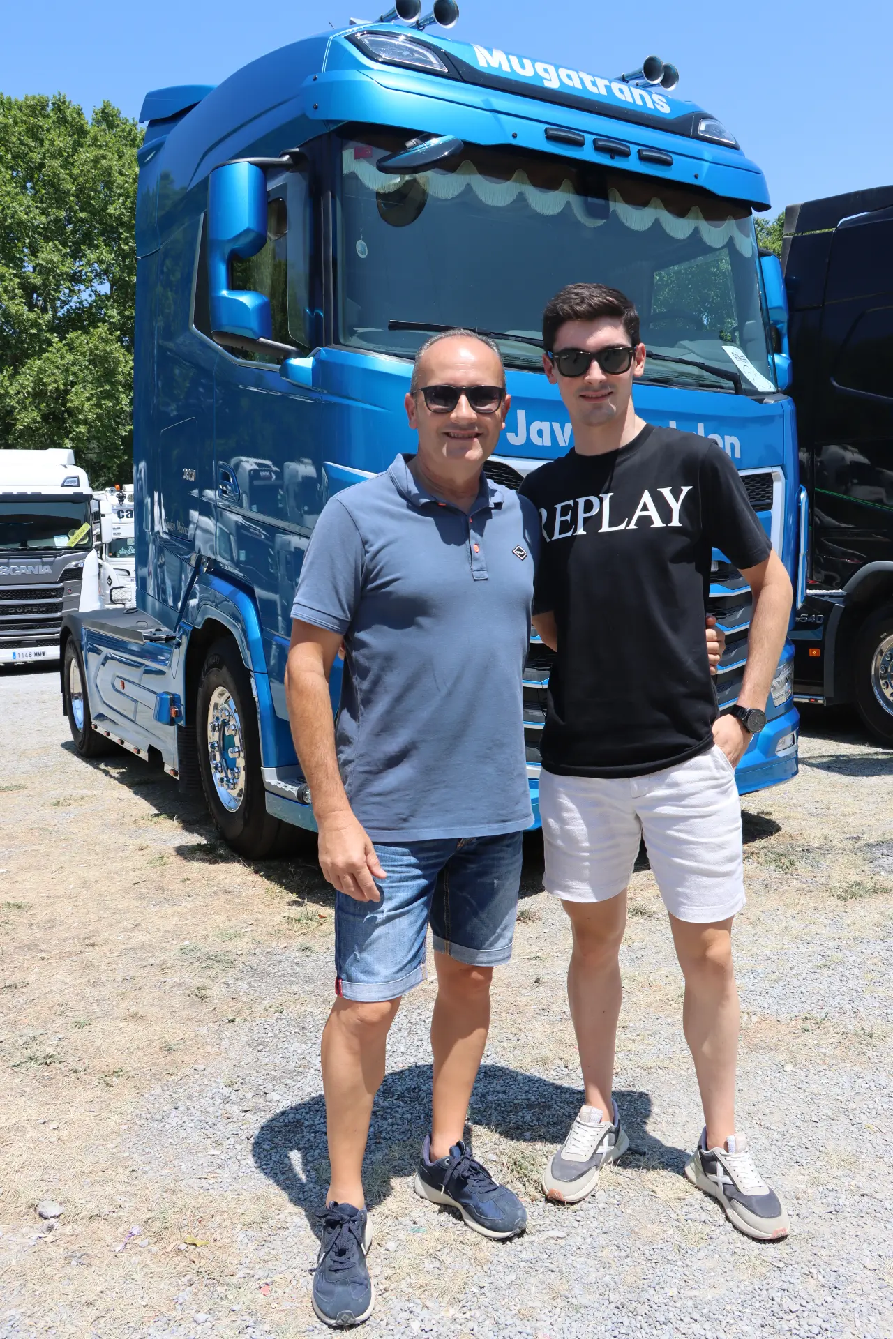 Dos hombres de pie delante de un gran camión azul del equipo en Olot, en un día soleado; ambos sonríen a la cámara con aire de concentración.