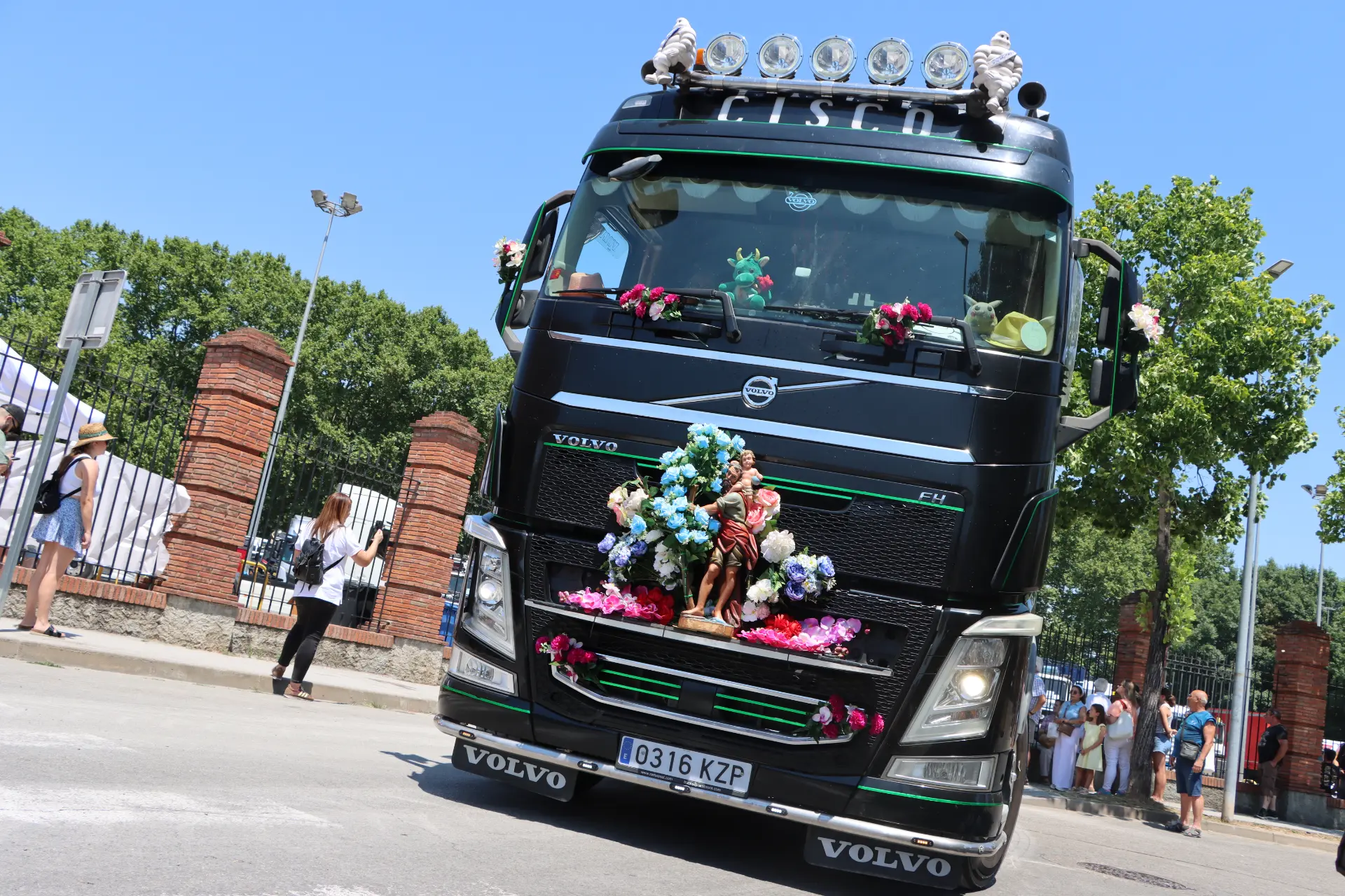 Camión Volvo negro decorado con flores y estatuas, circulando en un día soleado con gente al fondo durante la concentración de camiones de Olot, como un sincero homenaje a Oti Cabadas.