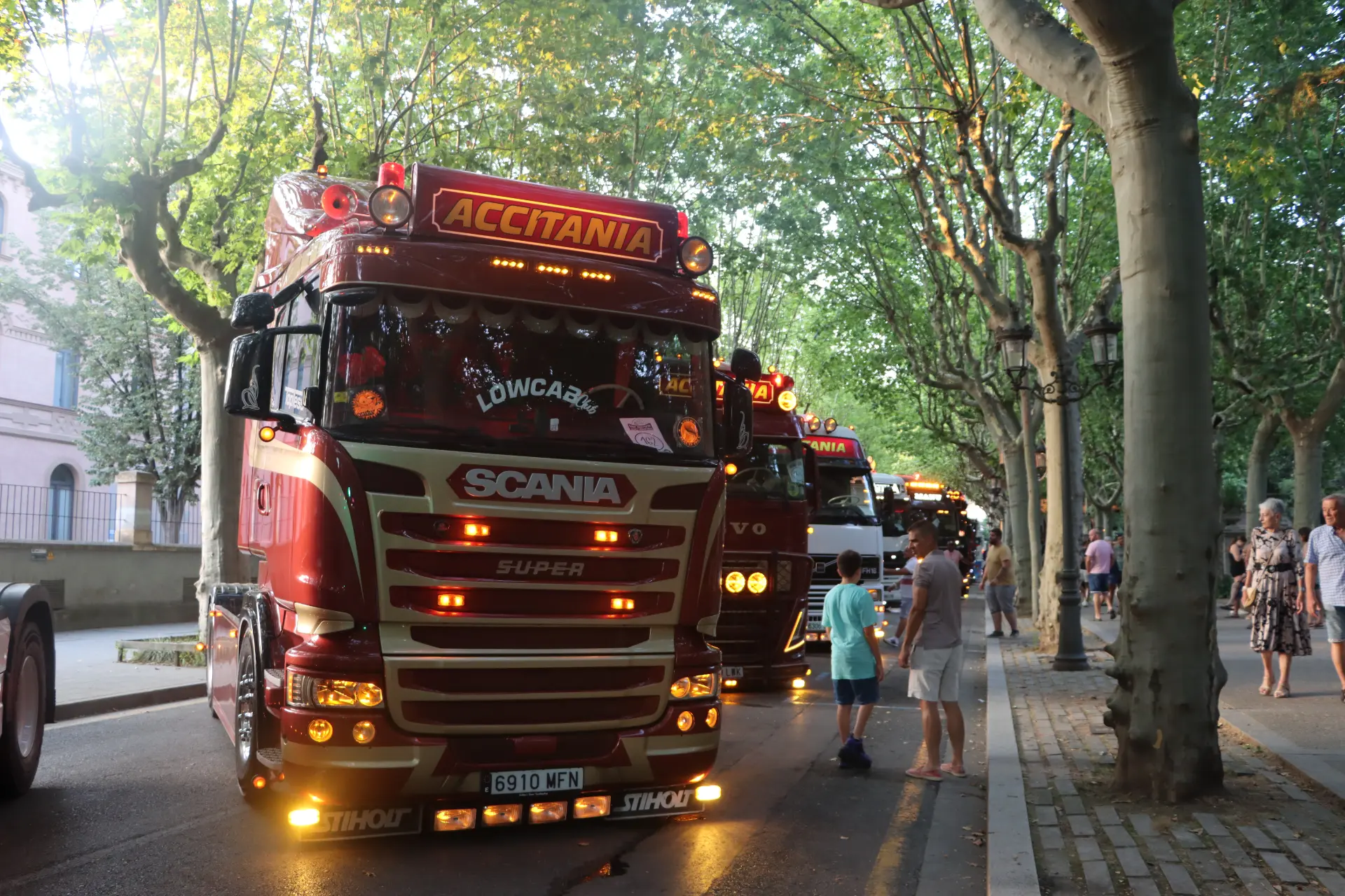 Los niños están de pie junto a unos camiones rojos aparcados con las luces encendidas, bajo los altos árboles que bordean una calle al atardecer, lo que evoca el animado ambiente de una concentración de camiones en Olot.