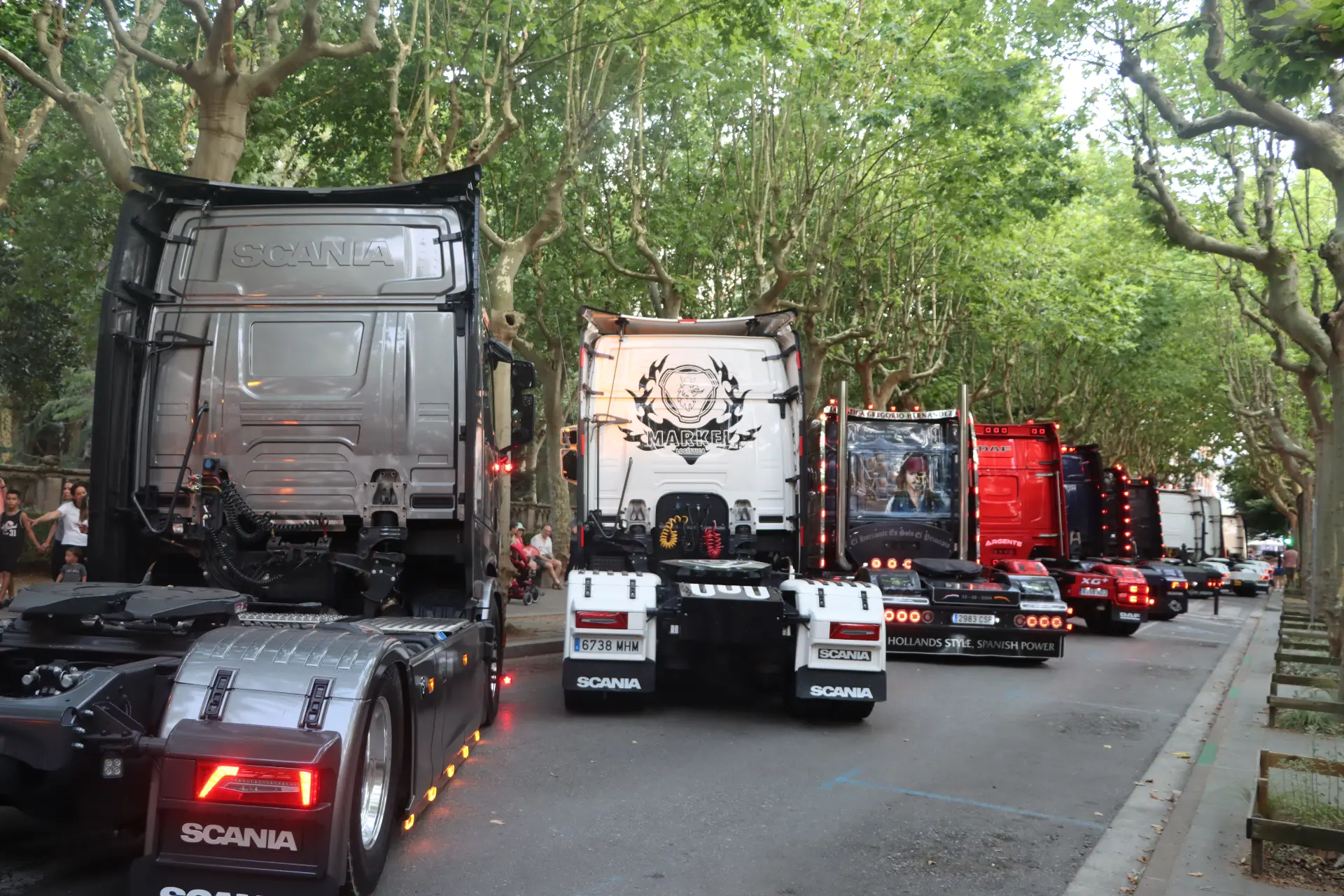 Varios camiones Scania decorados aparcados en una calle arbolada durante la concentración de camiones de Olot, con gente observando desde el lado izquierdo.