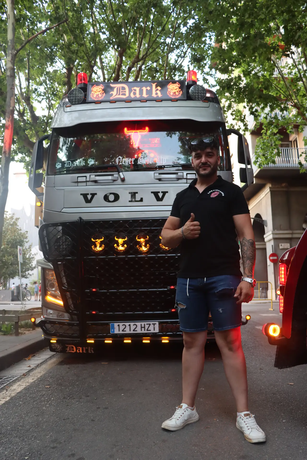 Un hombre con camiseta negra y pantalones cortos posa sonriendo y levantando los pulgares delante de un camión Volvo decorado en la calle, mostrando así su apoyo al equipo «Truck Olot».