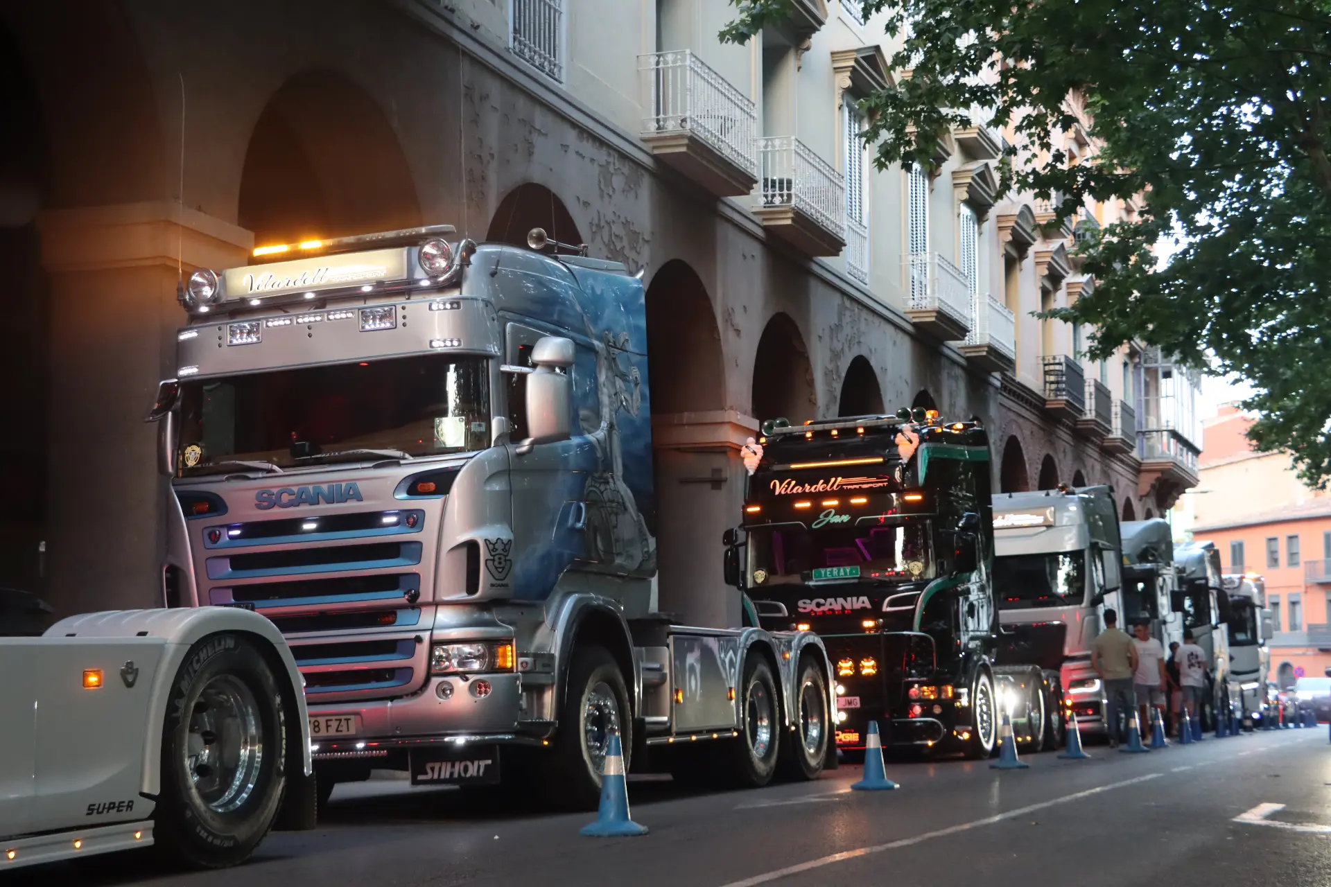 Unos camiones de colores aparcados en fila en una calle de la ciudad, junto a un edificio con arco y con árboles por encima, evocan el ambiente animado de una «concentración de camiones» en Olot.