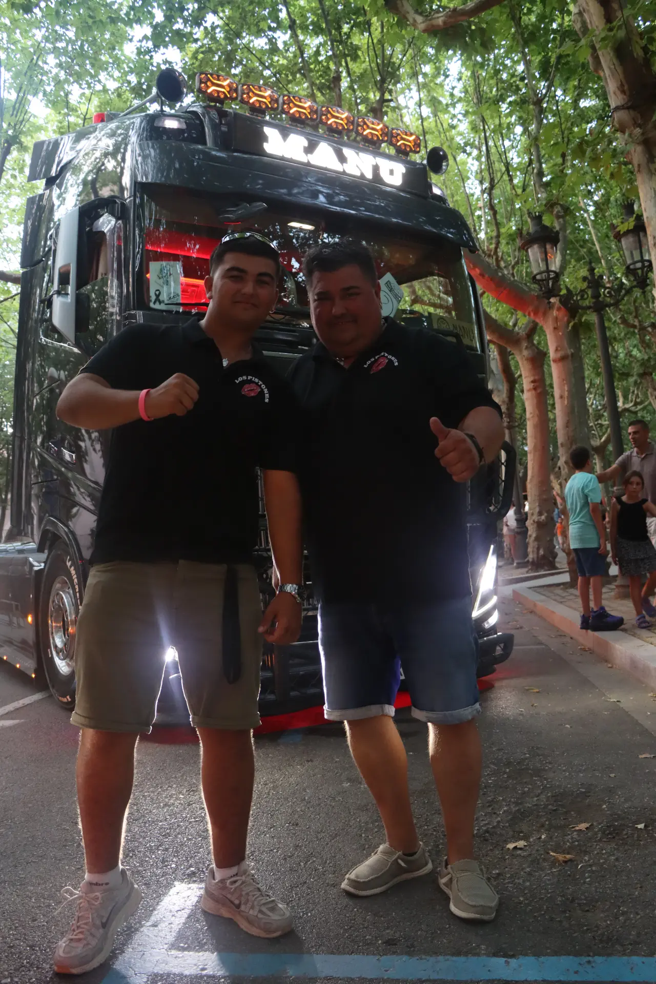Dos hombres posan sonrientes delante de una furgoneta negra en una calle arbolada, levantando el pulgar, representando con orgullo al equipo Truck Olot.