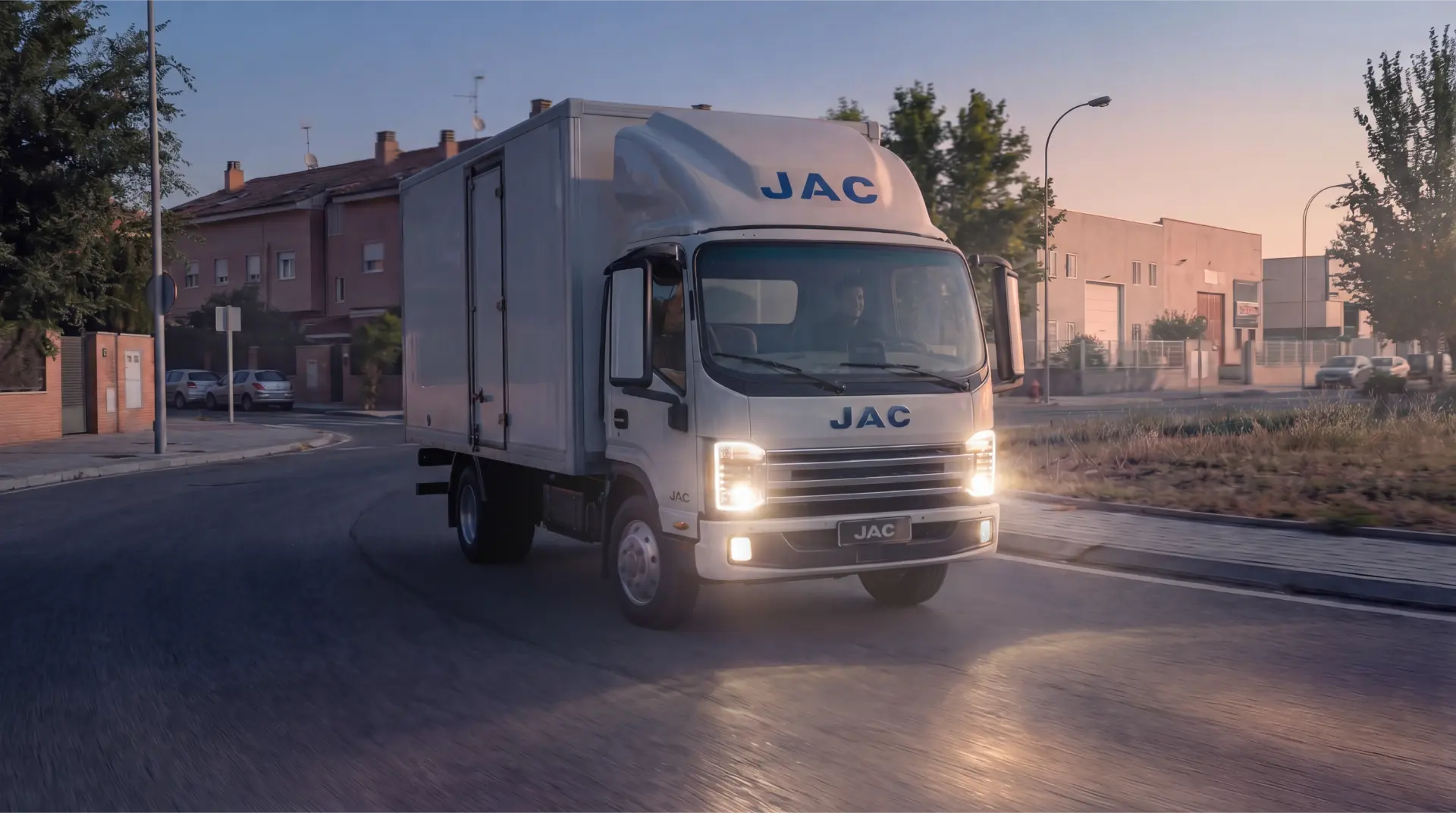 Una furgoneta de reparto JAC blanca que circula por una calle de las afueras al atardecer con los faros encendidos.