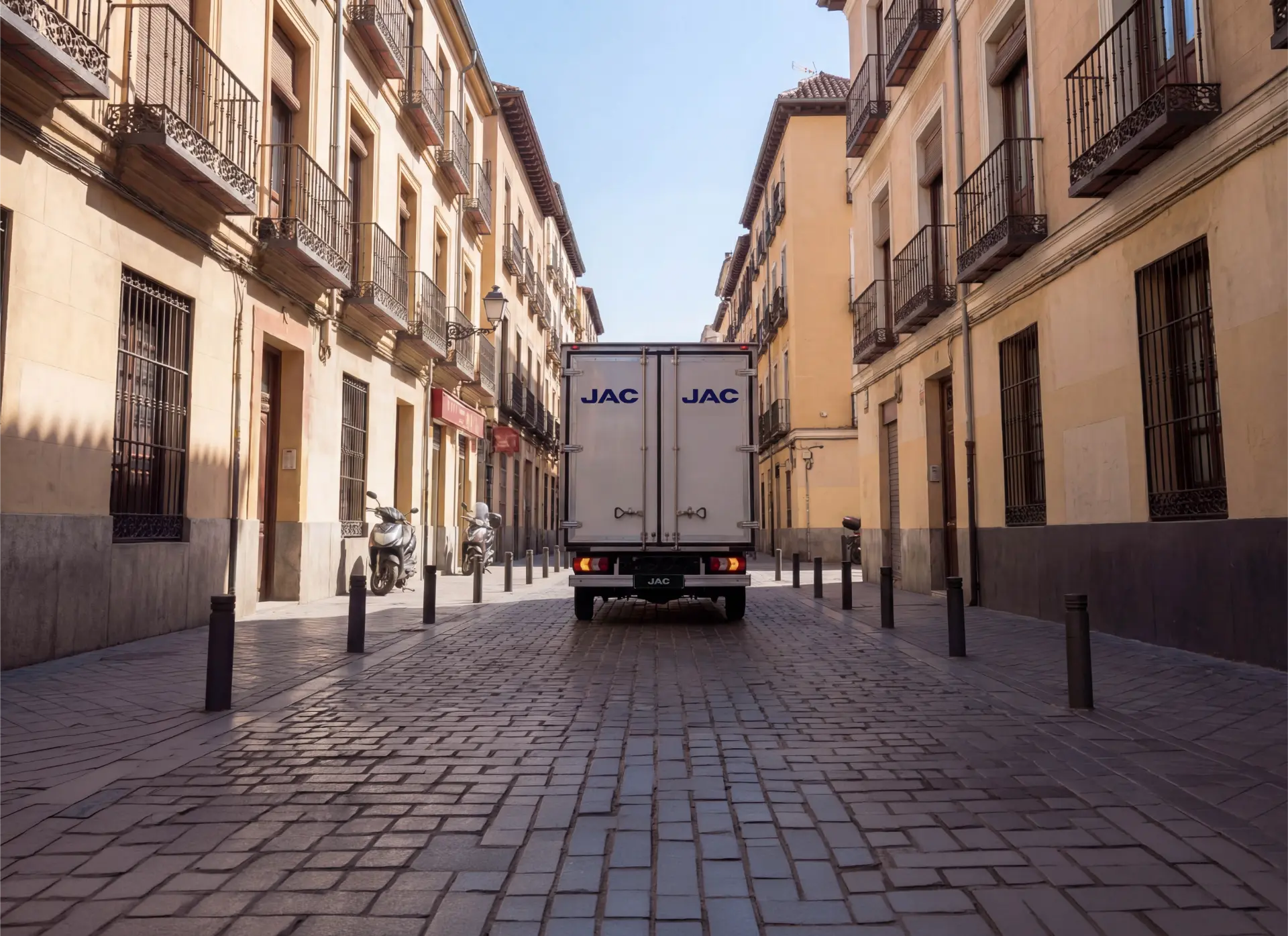 Una furgoneta de reparto blanca circula por una estrecha calle empedrada flanqueada por altos edificios amarillos.