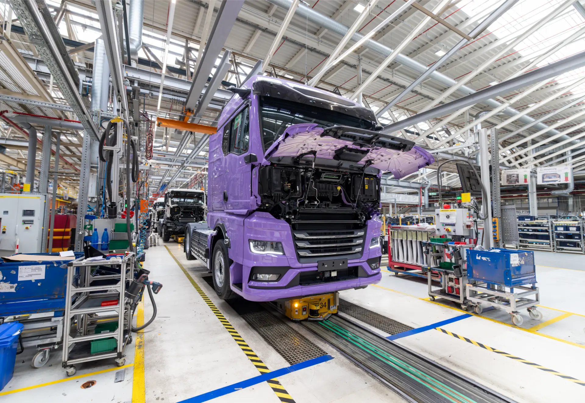 Un camión de color morado con la inscripción «Borrador automático» avanza a un ritmo constante por la cadena de montaje en el interior de una fábrica de automóviles muy bien iluminada.