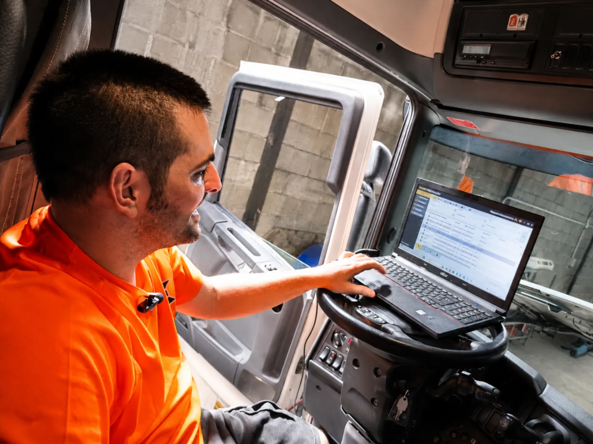 Un hombre con una camiseta naranja utiliza un ordenador portátil dentro de un vehículo con la puerta abierta, de cara a un muro de hormigón.