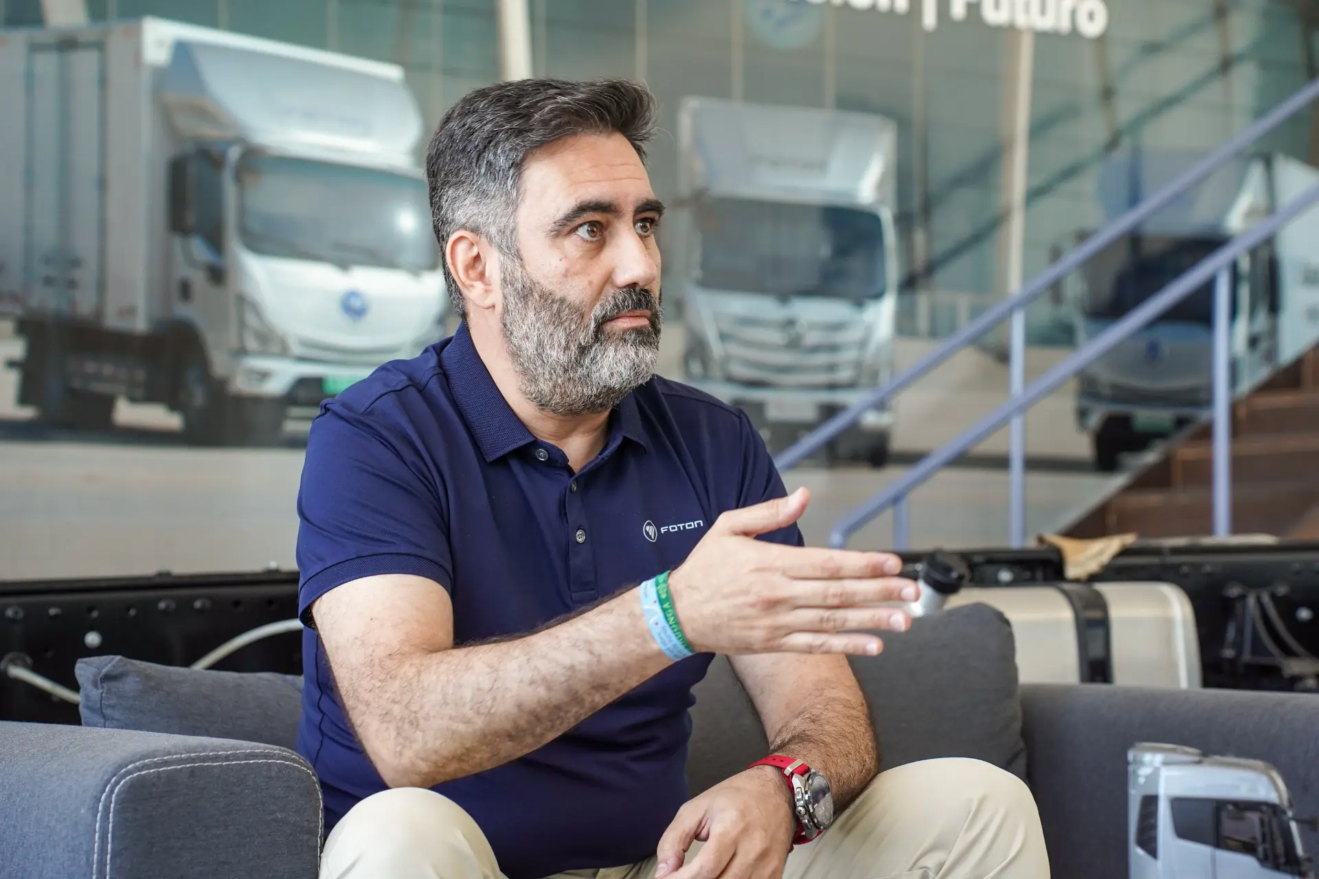 Un hombre con barba gesticula mientras está sentado en un sofá; al fondo se ven camiones de BMC.