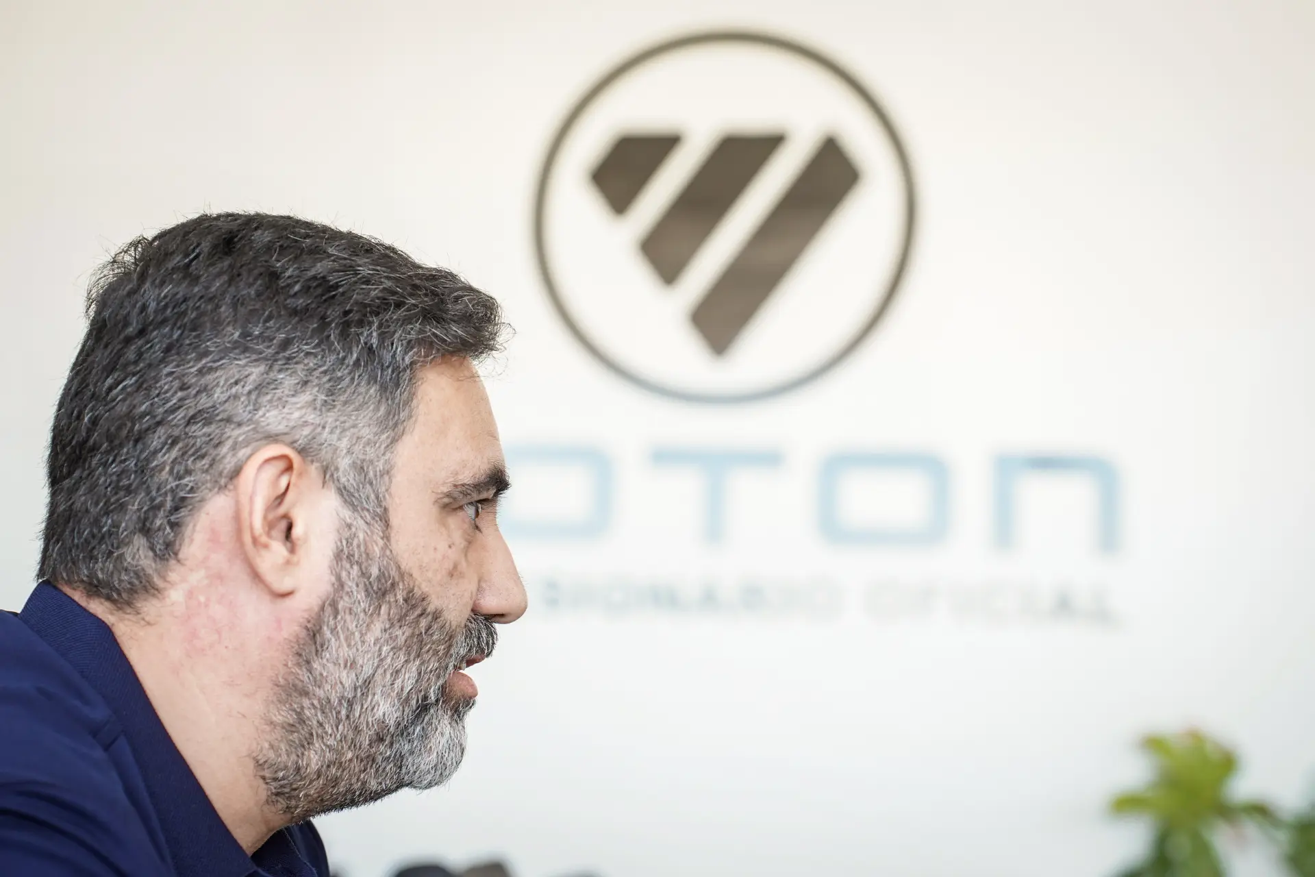 Un hombre de pelo y barba canosos, visto de perfil, está sentado junto a una pared en la que se ve un logotipo y un texto borrosos, posiblemente relacionados con «solo camión».