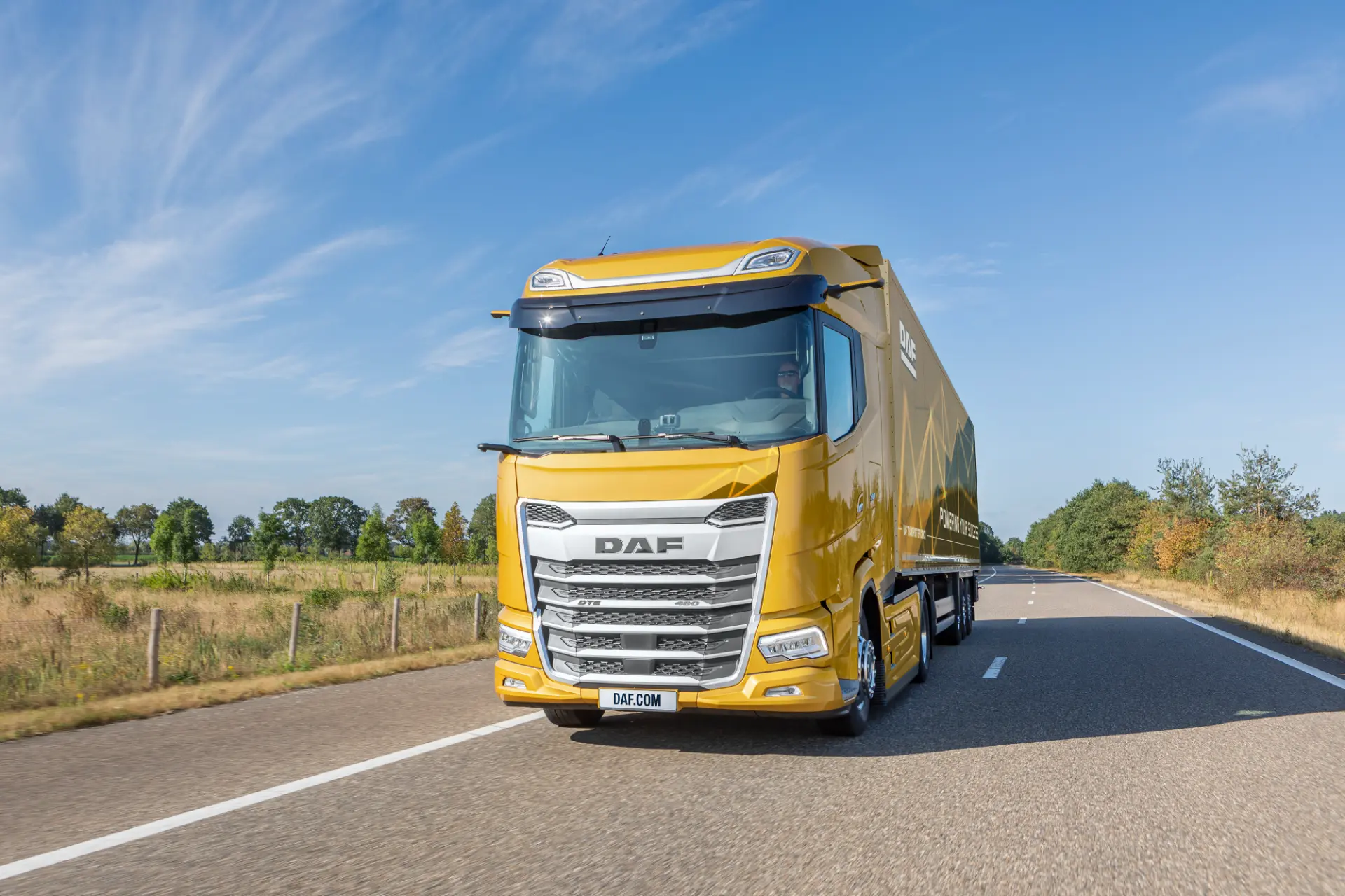 Camión eléctrico DAF XG+ de color amarillo circulando por una carretera rural, con el cielo azul y los árboles al fondo.