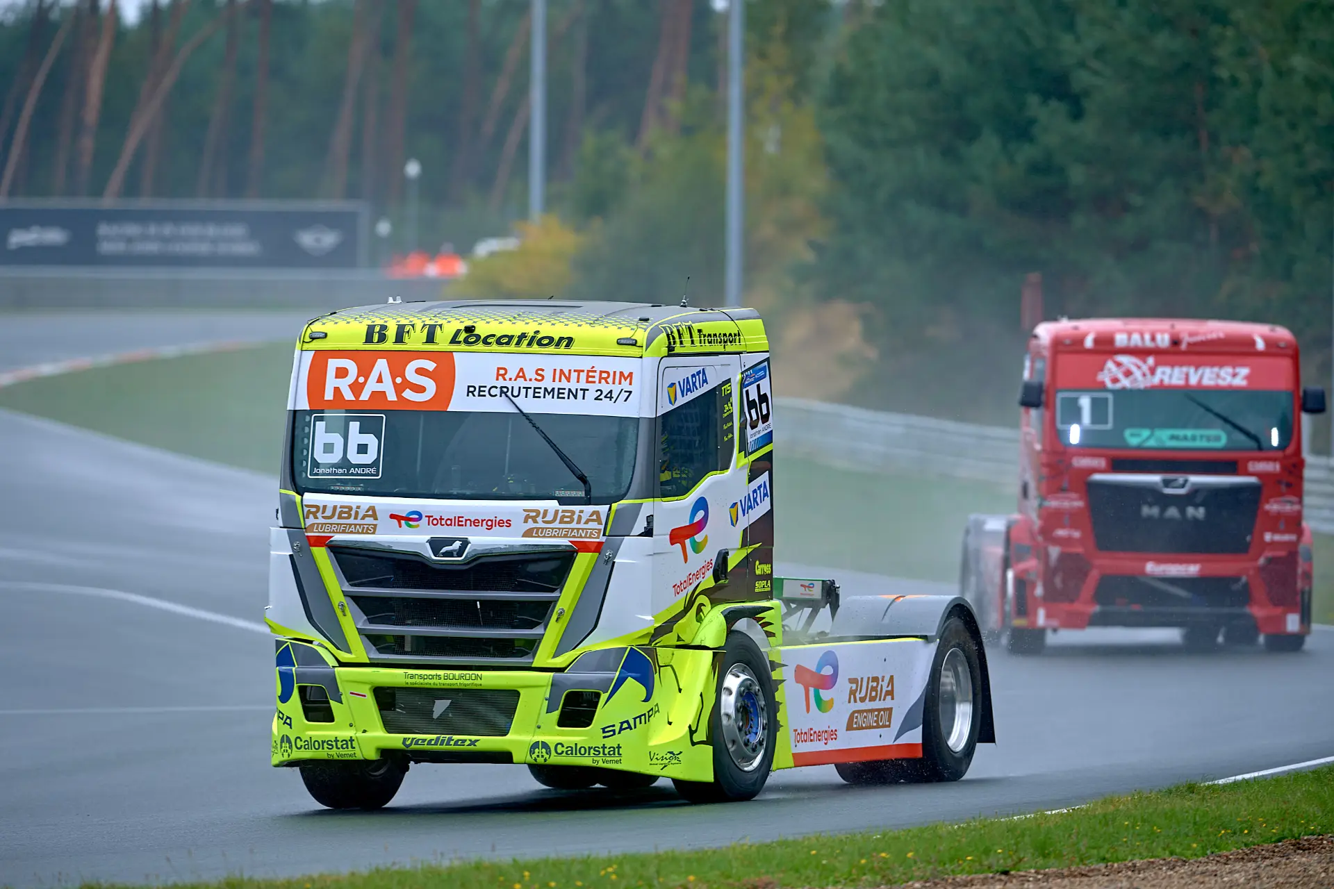 Dos camiones de carreras circulan por una pista mojada en Zolder, uno amarillo y blanco y el otro rojo, en un día nublado durante el Europeo de Camiones.