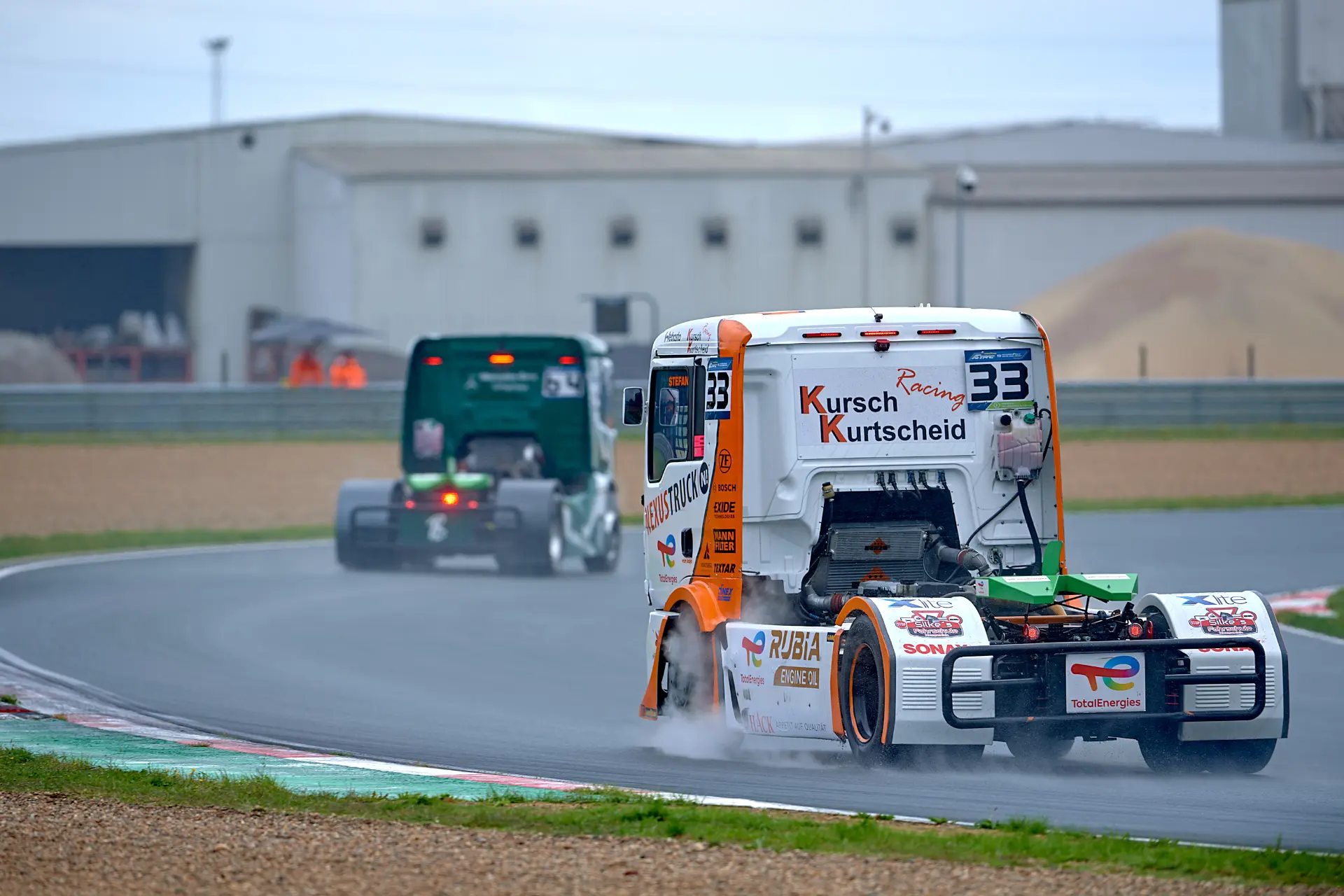 Dos camiones de carreras, entre ellos el potente Norbert Kiss, circulan por la pista mojada de Zolder, salpicando agua con los edificios de fondo, durante la emocionante prueba del Europeo de Camiones.