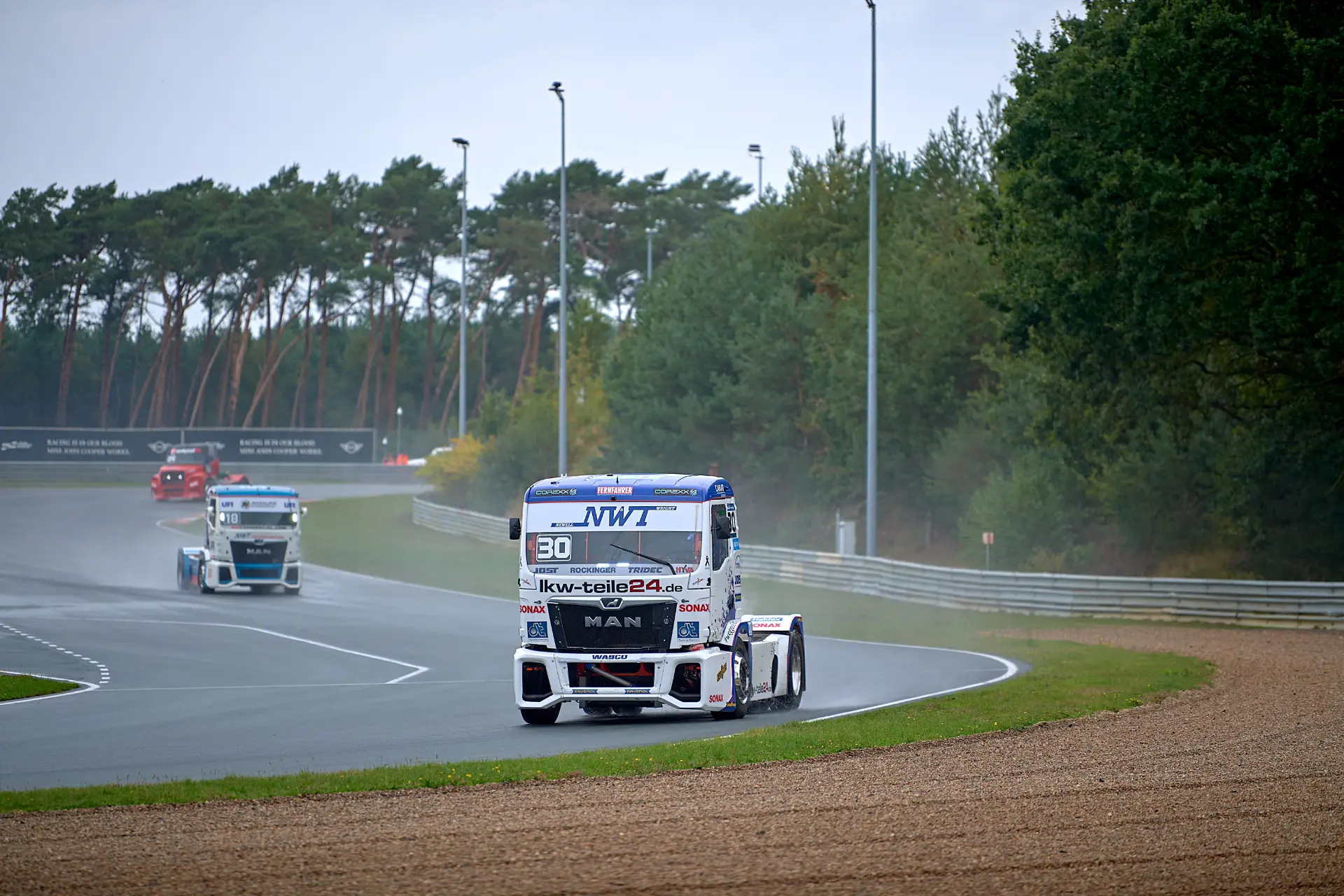 Tres camiones de carreras circulan por el circuito mojado de Zolder, rodeados de árboles y bajo un cielo nublado, mientras Norbert Kiss compite en el emocionante Campeonato Europeo de Camiones.