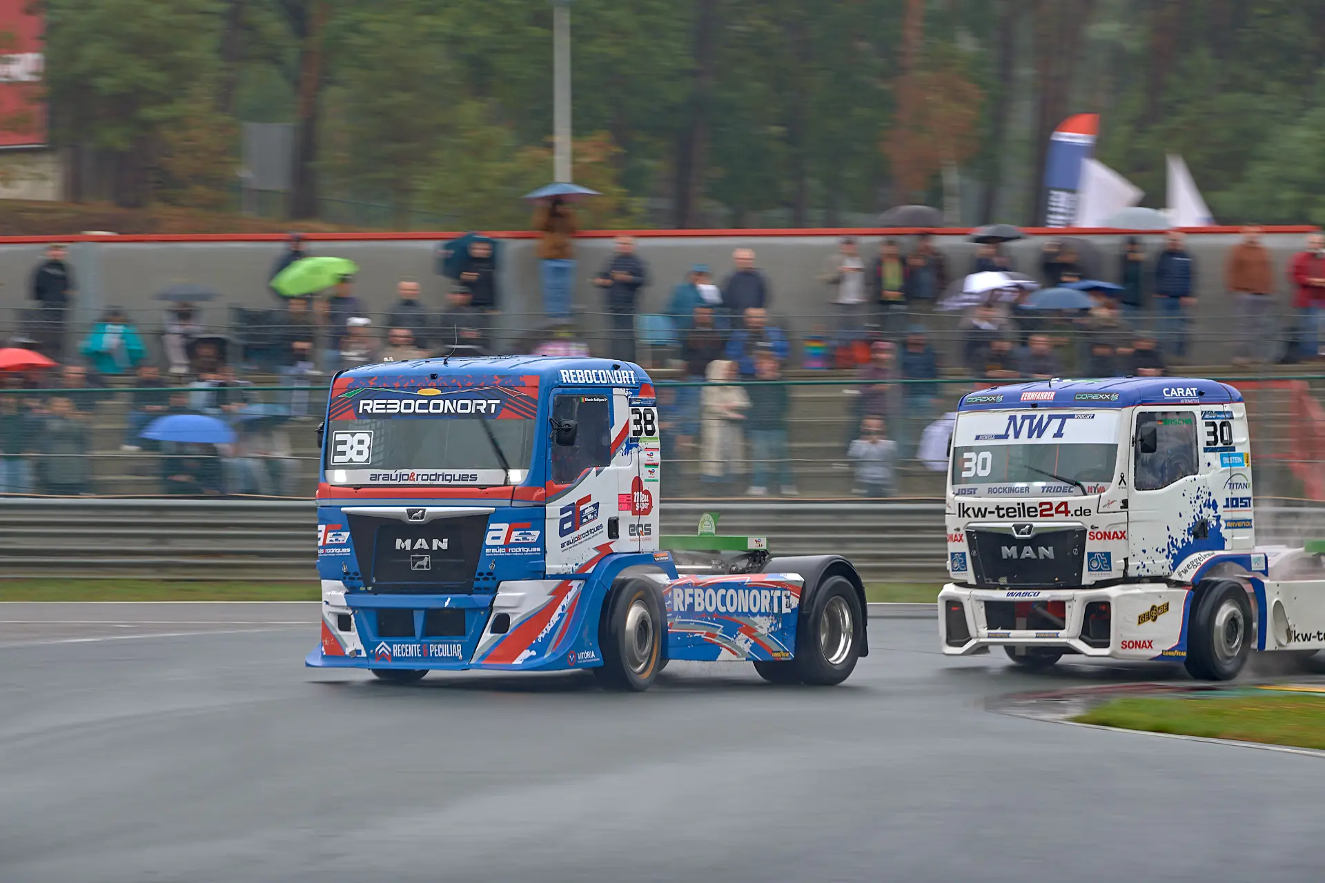 Dos camiones de carreras compiten en una pista mojada en Zolder, con los espectadores observando bajo los paraguas al fondo, durante la emocionante prueba del Europeo de Camiones, en la que participan pilotos de primer nivel como Norbert Kiss.