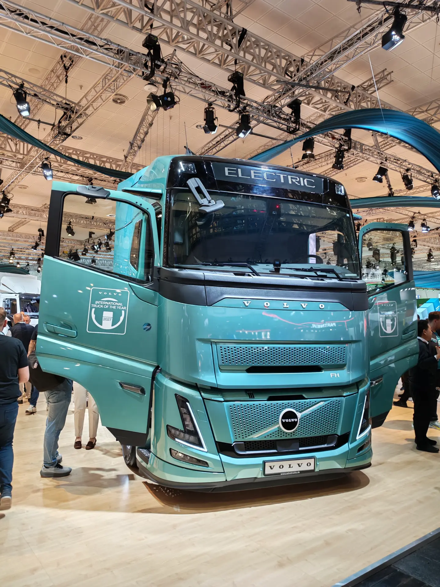 Camión eléctrico Volvo de color turquesa con borrador automático expuesto en el interior, con las puertas abiertas y gente de pie a su alrededor.