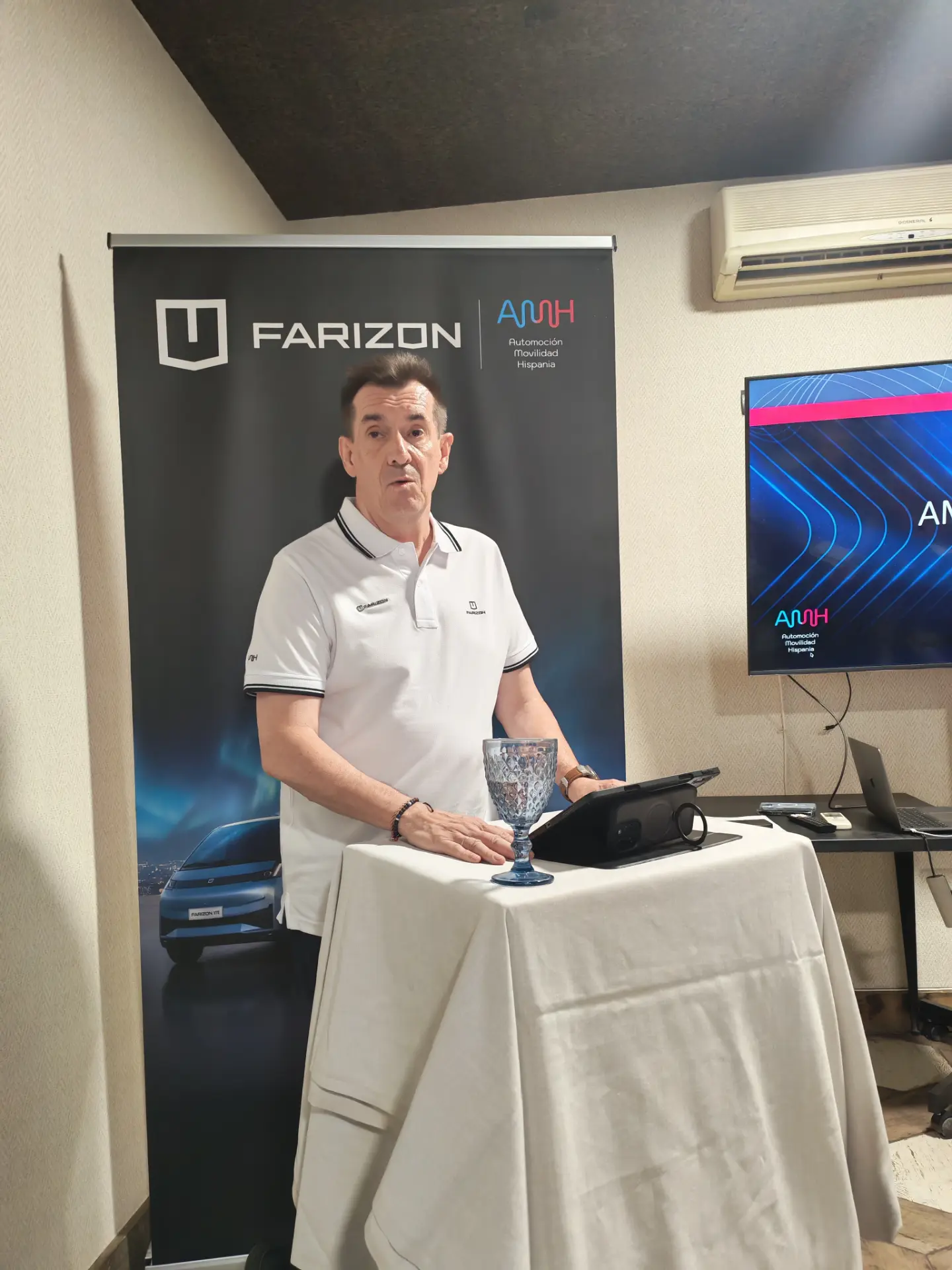 Un hombre vestido con un polo blanco habla desde un atril con una pancarta de Farizon y una pantalla de televisión al fondo, en la que se muestran las innovadoras furgonetas eléctricas Farizon SV y V7E.