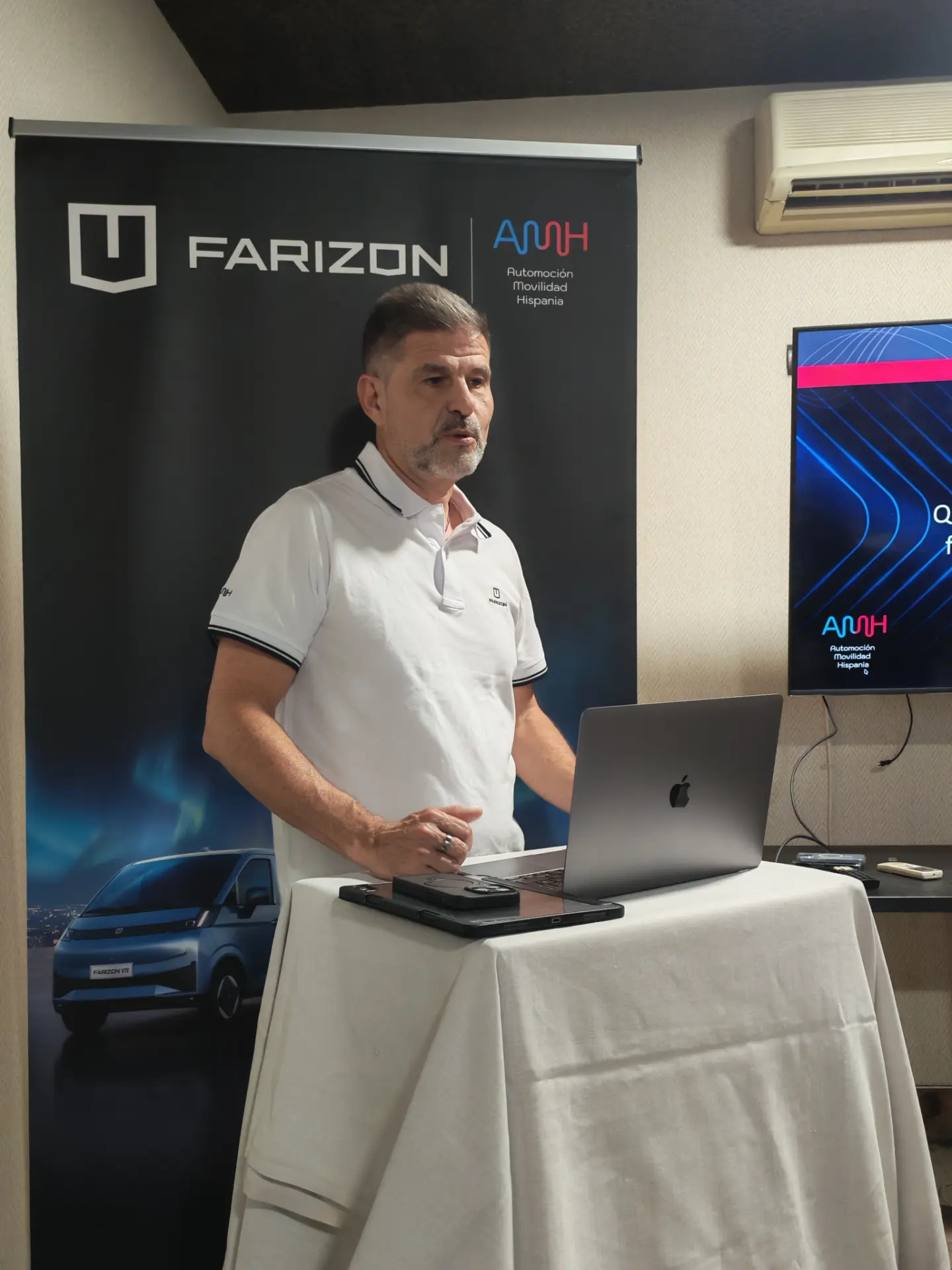 Un hombre con camisa blanca aparece en un estrado con un ordenador portátil, de pie junto a un cartel de Farizon y una pantalla en la que se muestran las innovadoras furgonetas eléctricas Farizon SV y V7E.