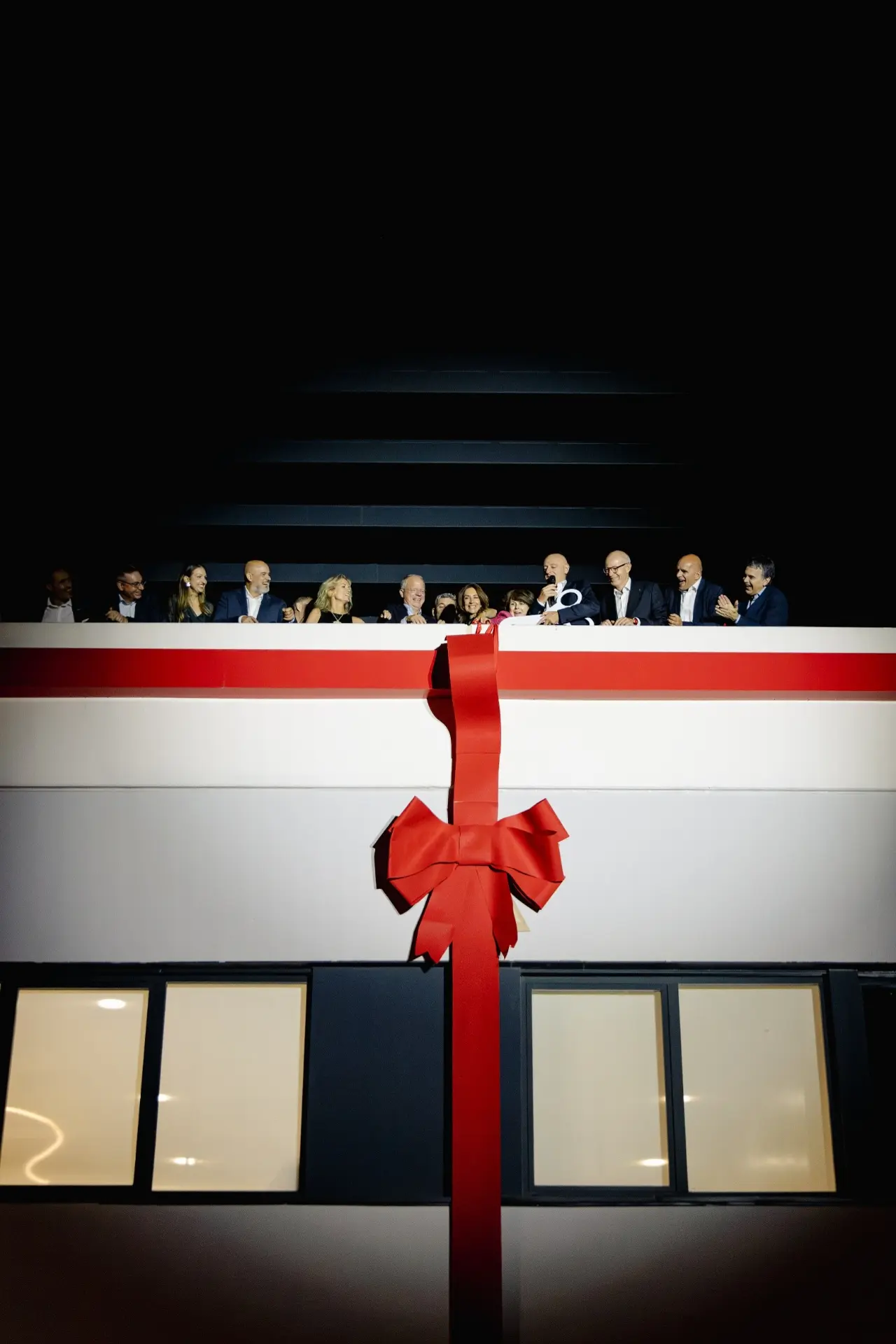 Varias personas se encuentran en un balcón situado en lo alto de un edificio decorado con una gran cinta roja y un lazo, por la noche, celebrando la nueva sede de Renault Trucks en el Truck Center Madrid.
