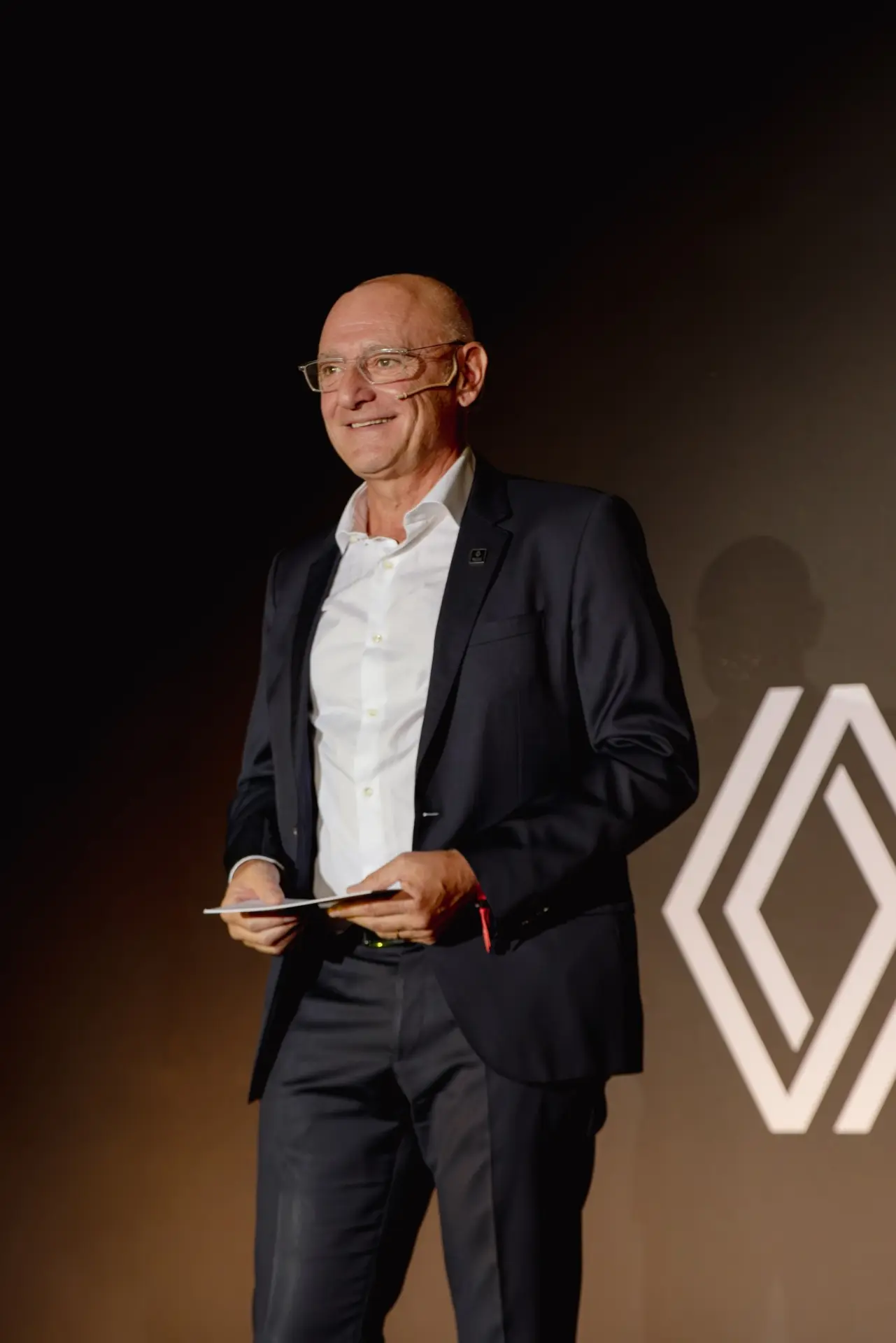 Un hombre vestido con traje y con unos documentos en la mano posa frente a un logotipo blanco, moderno y geométrico sobre un fondo oscuro, que representa la nueva sede de Renault Trucks en el Truck Center Madrid, en Getafe.