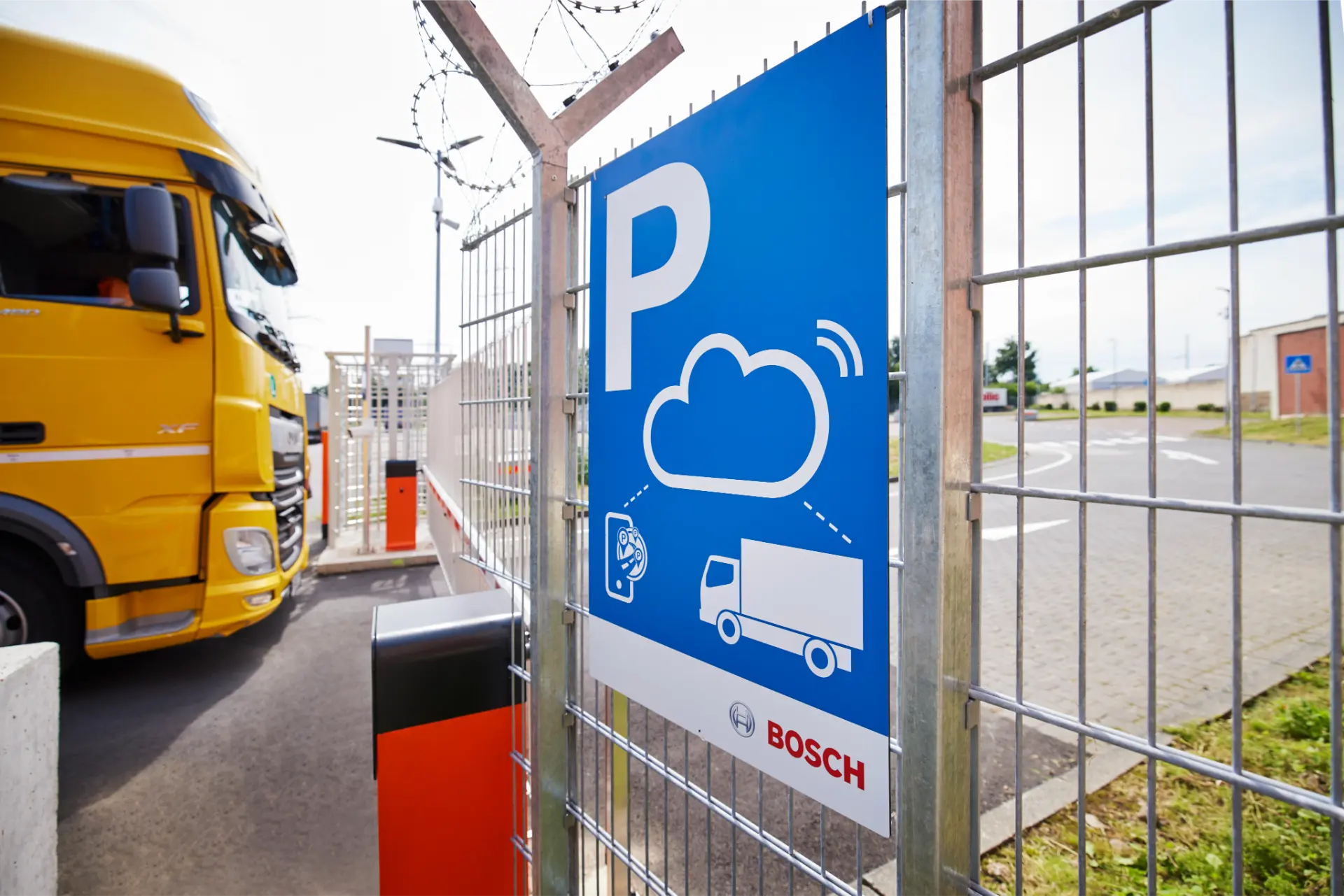 Señal de aparcamiento azul para camiones con el símbolo de una nube, que indica «borrador automático», junto a un camión amarillo en un aparcamiento cerrado.