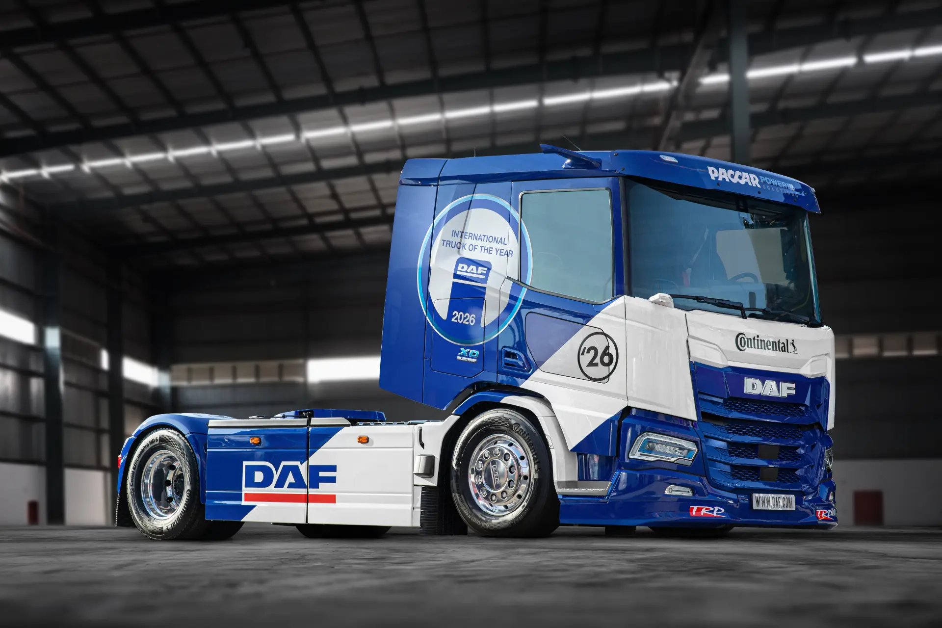 Cabina de camión DAF azul y blanca con el número 26, aparcada en el interior de una gran nave industrial, que recuerda a un camión de carreras DAF o a la innovadora serie XD Electric, ideal para tareas como el transporte de leche.