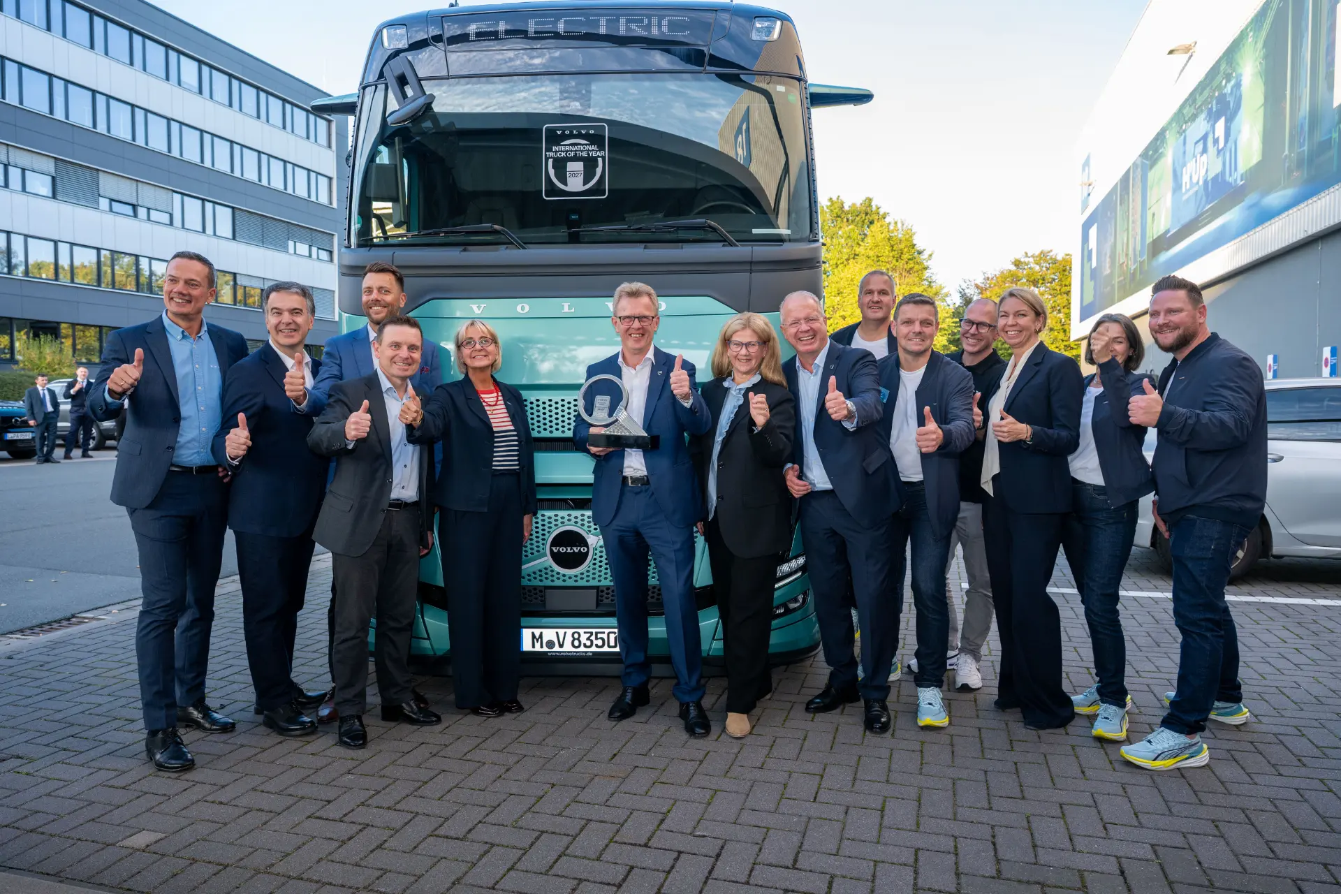 Un grupo de personas vestidas con traje de negocios posa sonriente y levantando el pulgar delante de un gran camión de color verde azulado, celebrando la integración satisfactoria de un sistema de borrador automático en sus operaciones.