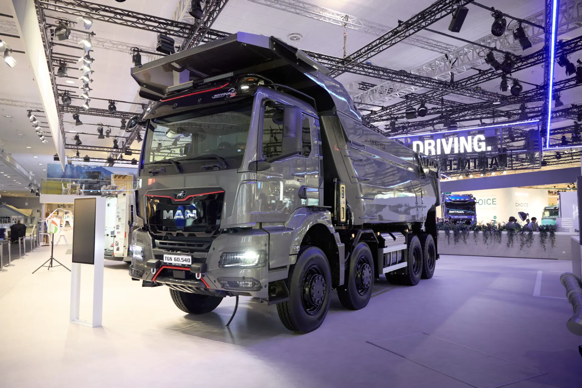 Un gran camión volquete MAN eTGX de color gris expuesto en el interior de una feria comercial, en el que se destacan su impresionante autonomía de 720 kilómetros y su capacidad de carga rápida, con luces y elementos expositivos al fondo.