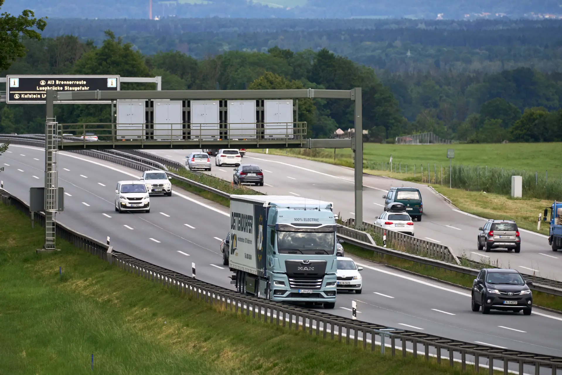 Varios coches y un camión circulan por una autopista con carriles separados, con vegetación y árboles al fondo, lo que ilustra los esfuerzos continuos en pro de la descarbonización impulsados por la Iniciativa Brennero Verde a lo largo de la ruta del Brennero.