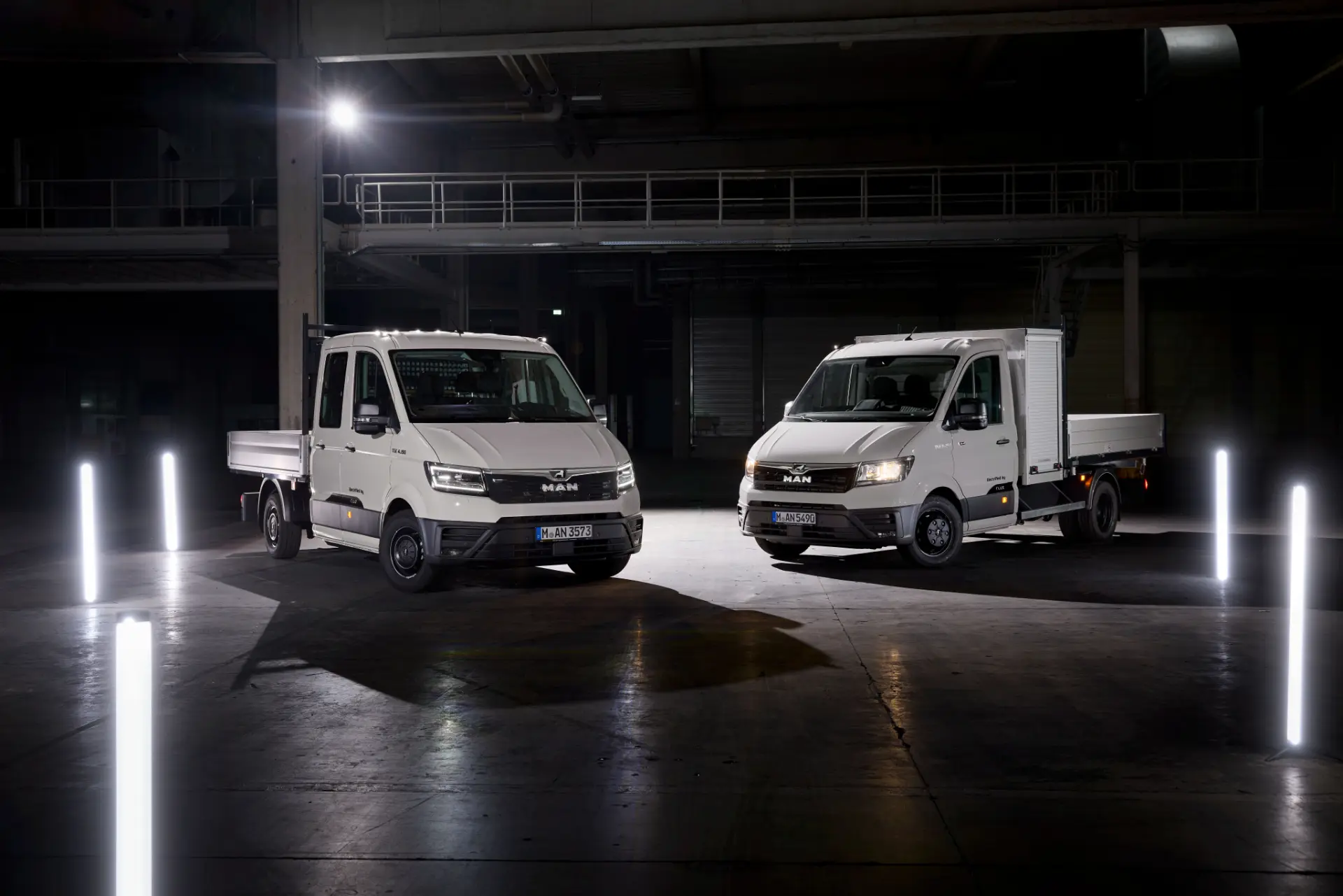 Dos camiones comerciales MAN eTGX de plataforma, de color blanco, equipados con capacidad de carga rápida y una impresionante autonomía de 720 kilómetros, están aparcados en una nave industrial con poca luz y llamativas luces verticales.