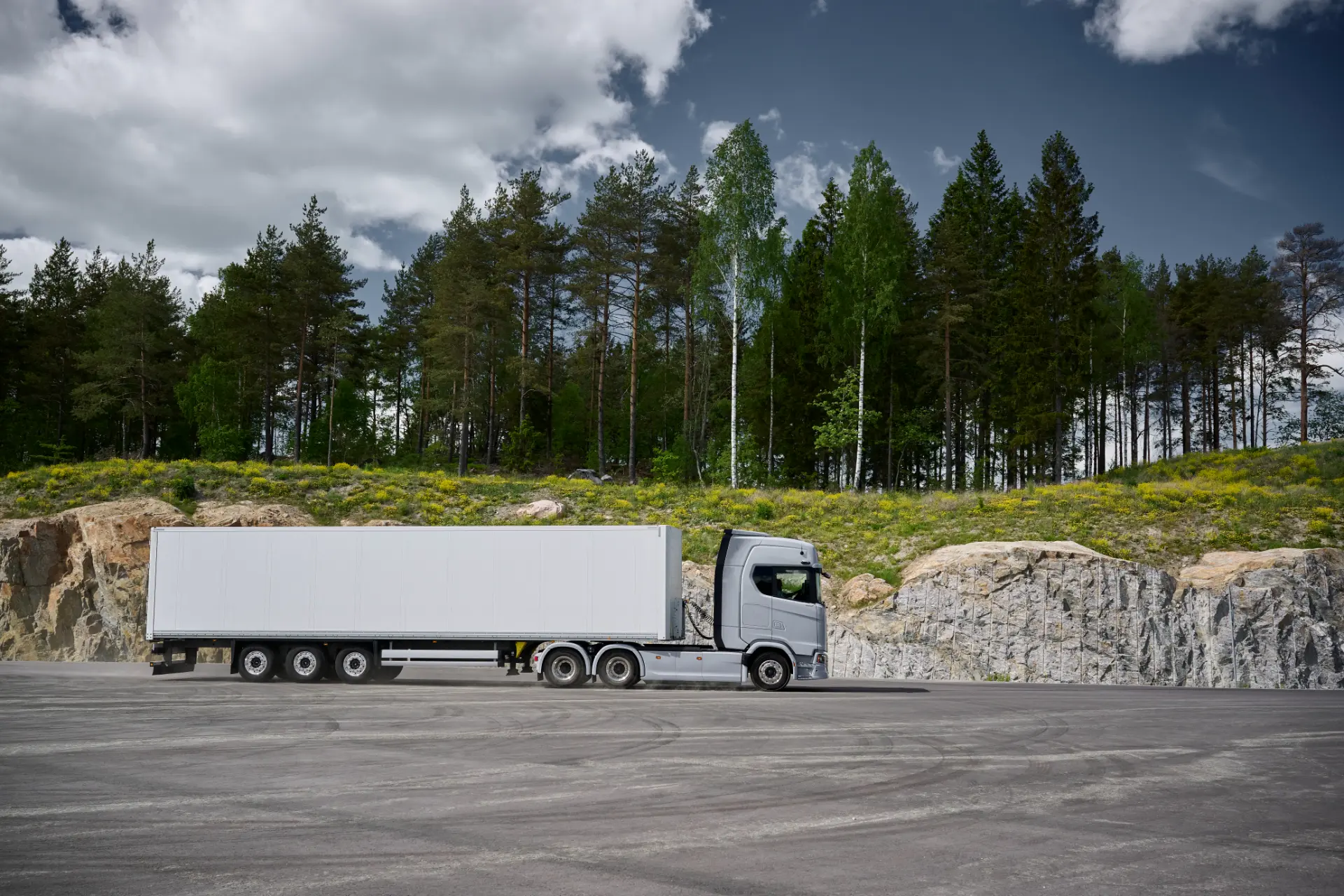 Un camión articulado plateado con un remolque blanco aparcado junto a un bosque, como si transportara una lijadora eléctrica.