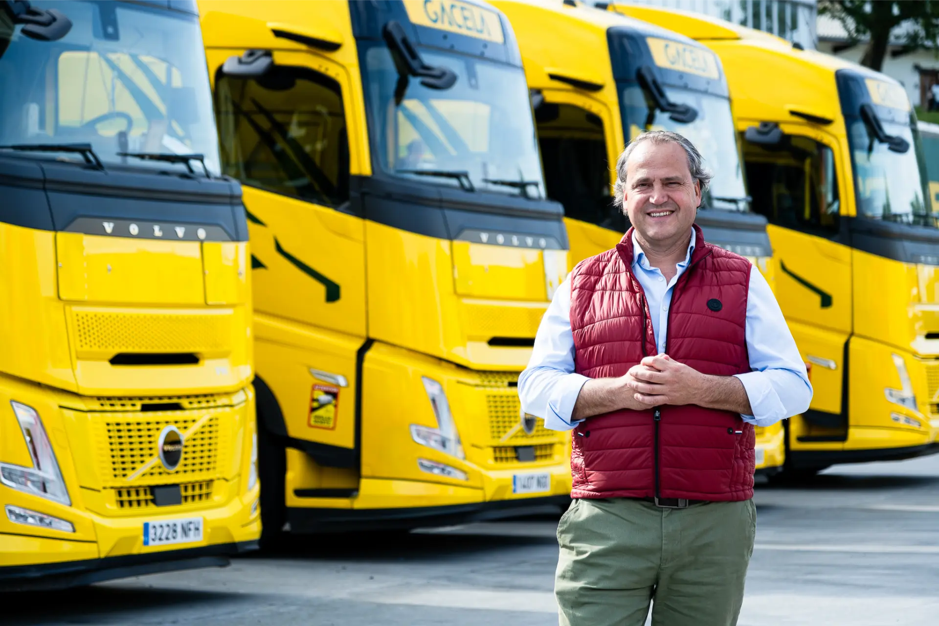 Un hombre con un chaleco rojo está de pie delante de varios camiones Volvo FH Aero de Transportes Gacela.
