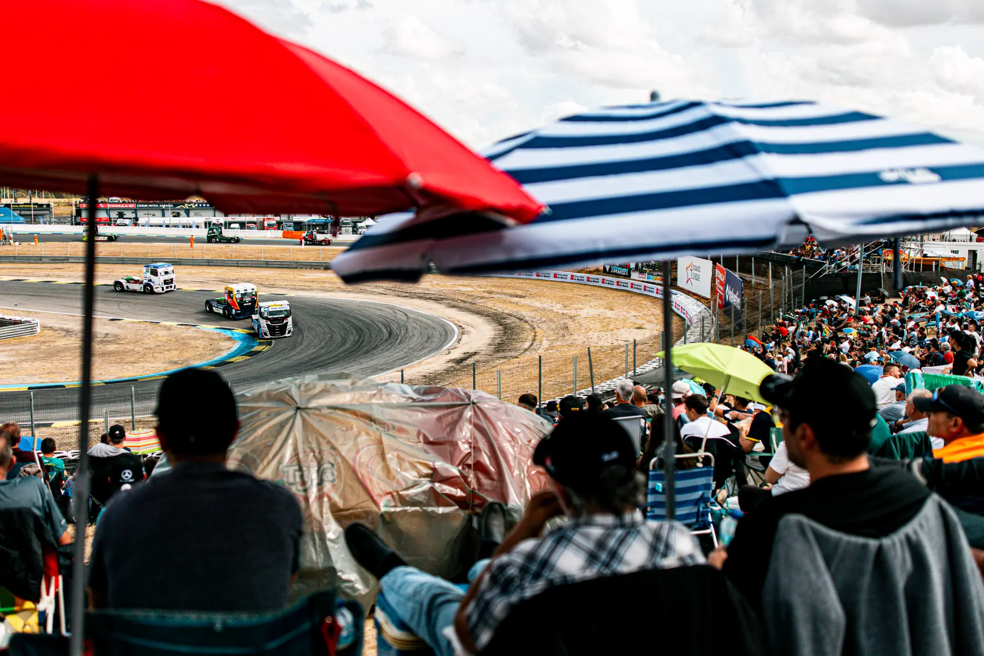 Los espectadores, bajo sus paraguas, observan cómo los camiones toman a toda velocidad una curva en el Jarama Europeo de Camiones.