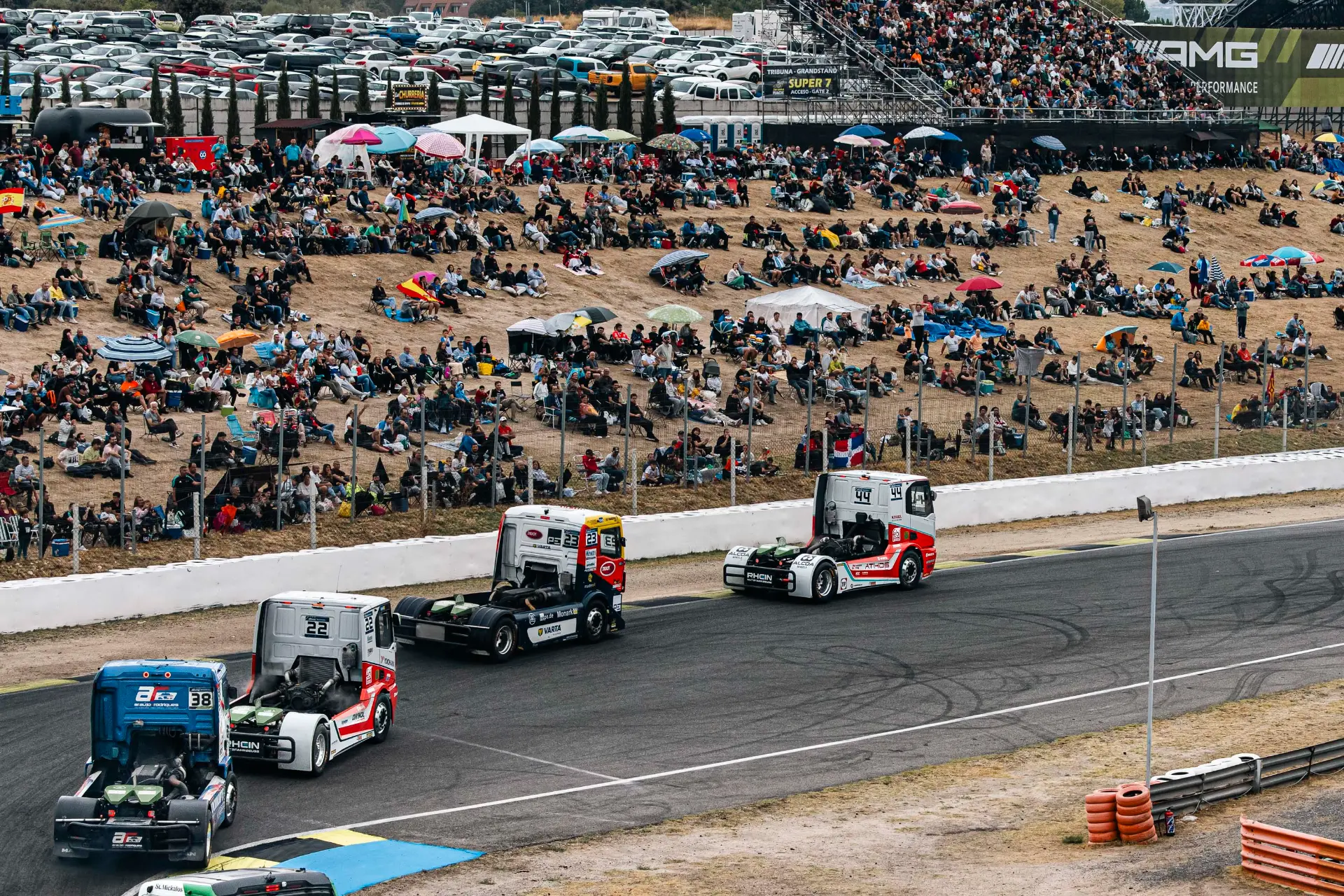 Tres camiones articulados, entre ellos el de Norbert Kiss, compiten en el Circuito del Jarama ante una multitud reunida en una colina cubierta de hierba.